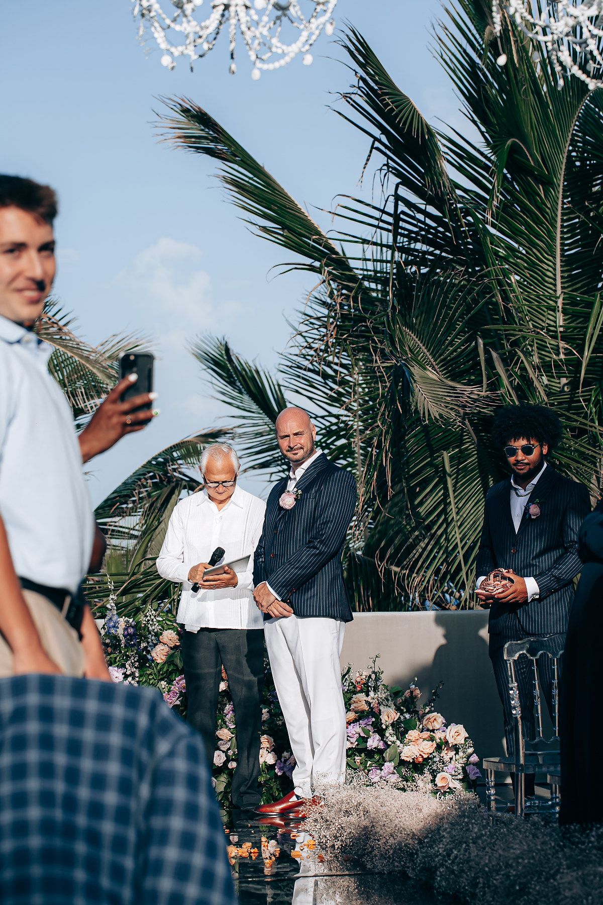 An outdoor wedding ceremony scene with palm trees and floral decorations in the background.