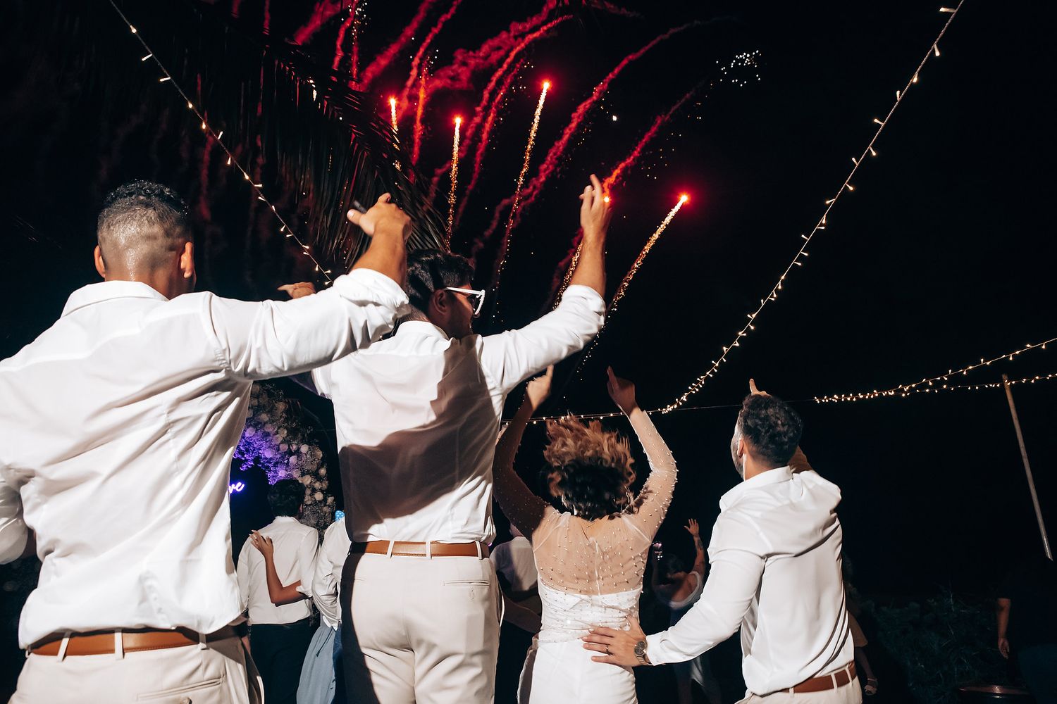 Wedding party in white attire dancing together with arms raised against a dark background with red lighting effects.