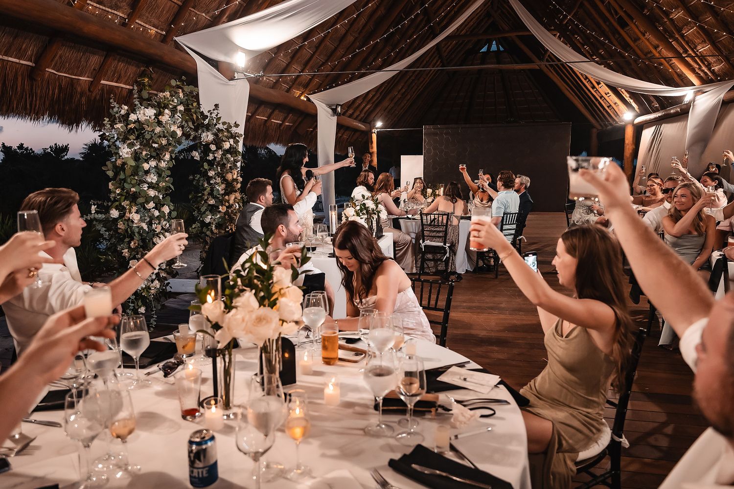 Wedding reception guests raise their glasses in celebration inside a rustic barn venue decorated with twinkling lights.