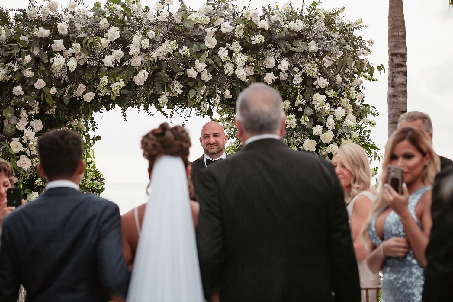 An outdoor wedding ceremony takes place under a beautiful floral arch adorned with white blooms and greenery.
