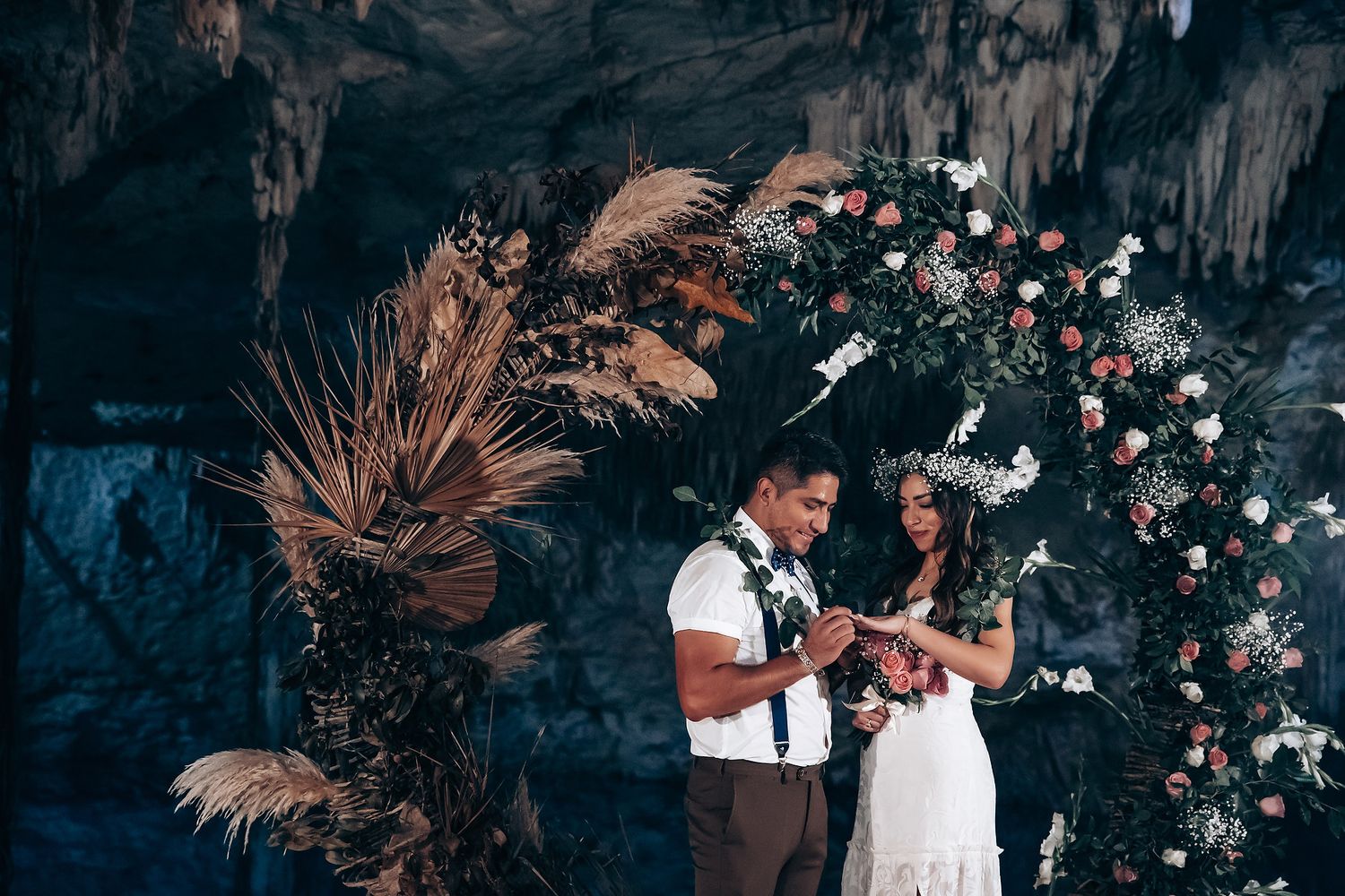 Romantic wedding ceremony under floral arch with pampas grass and roses against dramatic cave backdrop.