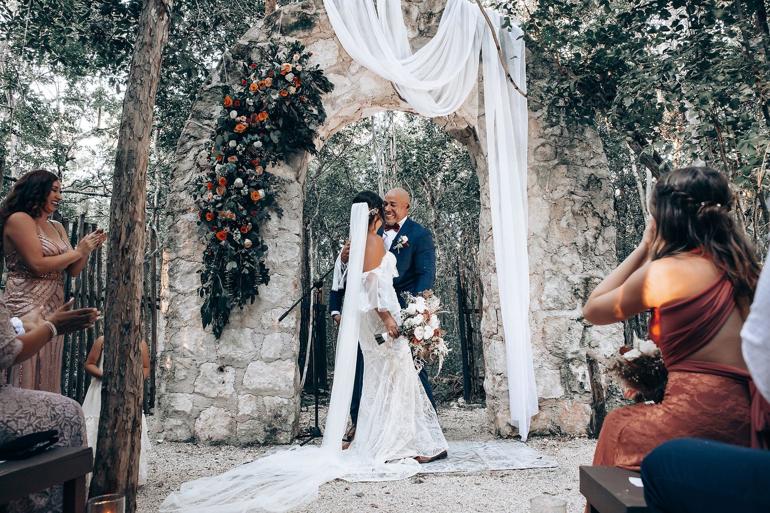 Outdoor wedding celebration with guests gathered under trees and white fabric decorations.