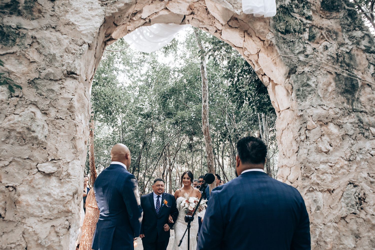 A wedding ceremony takes place under a rustic stone archway surrounded by trees and greenery.