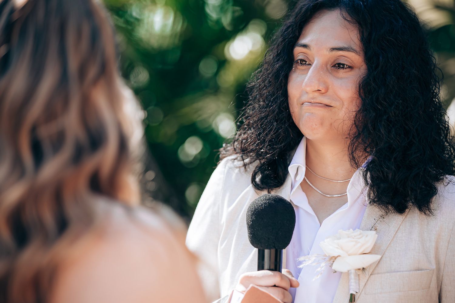 A person with curly dark hair wearing a white blouse smiles and gestures while speaking in an outdoor setting.