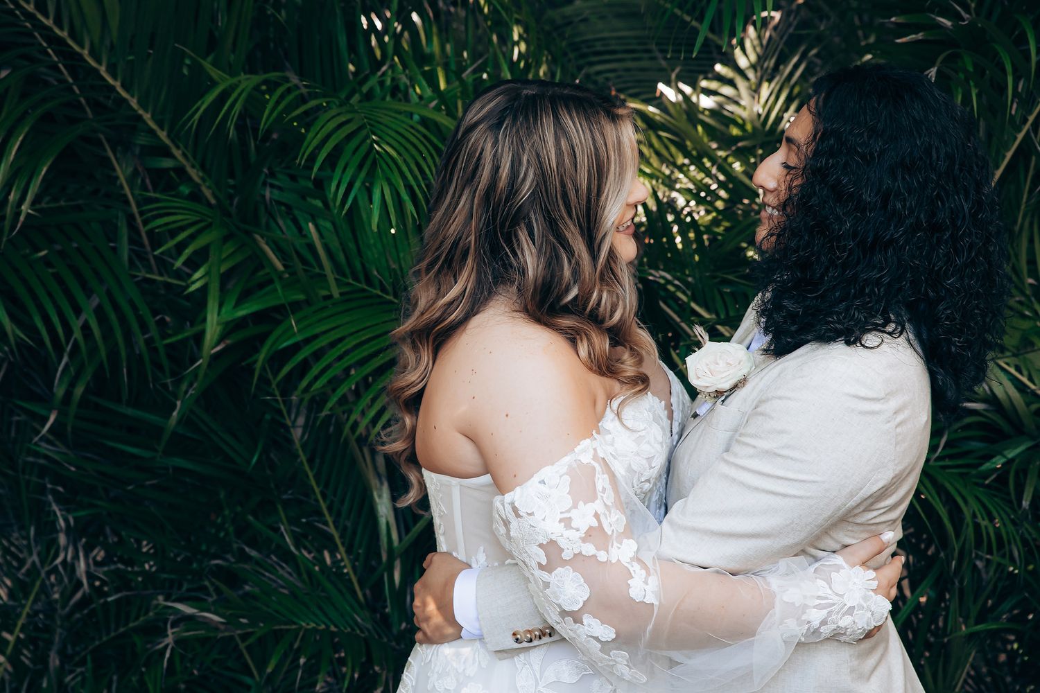 A couple in wedding attire embraces romantically in a lush garden setting with palm trees in the background.