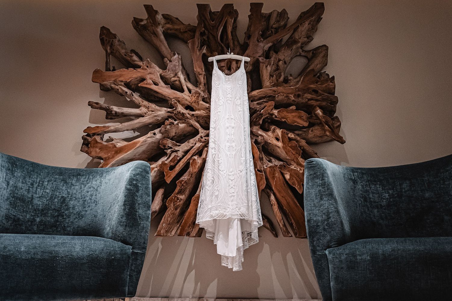 White wedding gown suspended beneath wooden sunburst chandelier with blue velvet sofa visible below.
