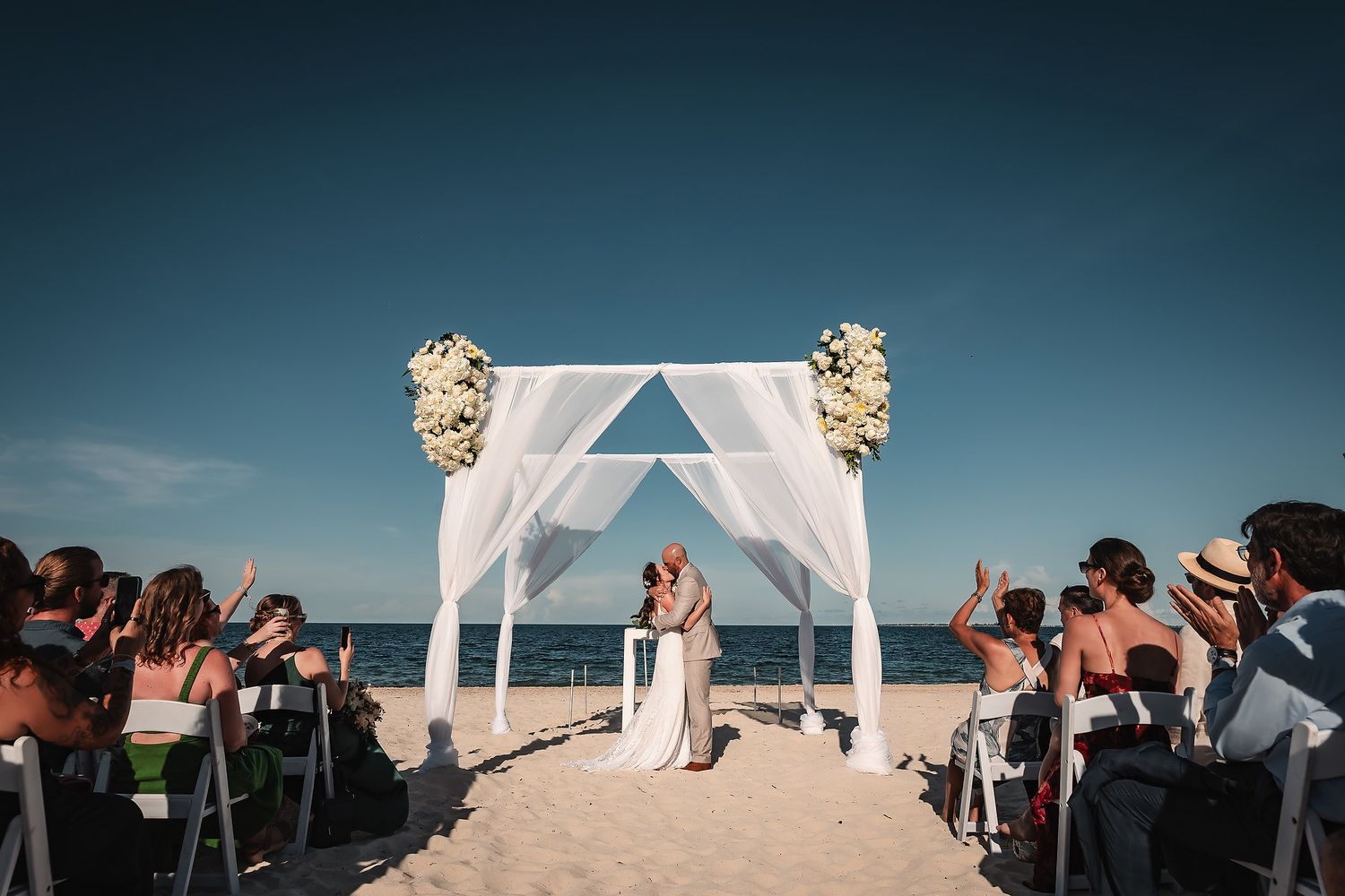 Wedding ceremony on the beach with white arch, floral decorations, and guests seated on either side.