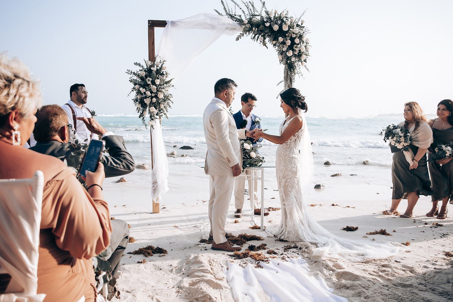 A beachfront wedding ceremony under a floral arch with guests watching from white chairs.