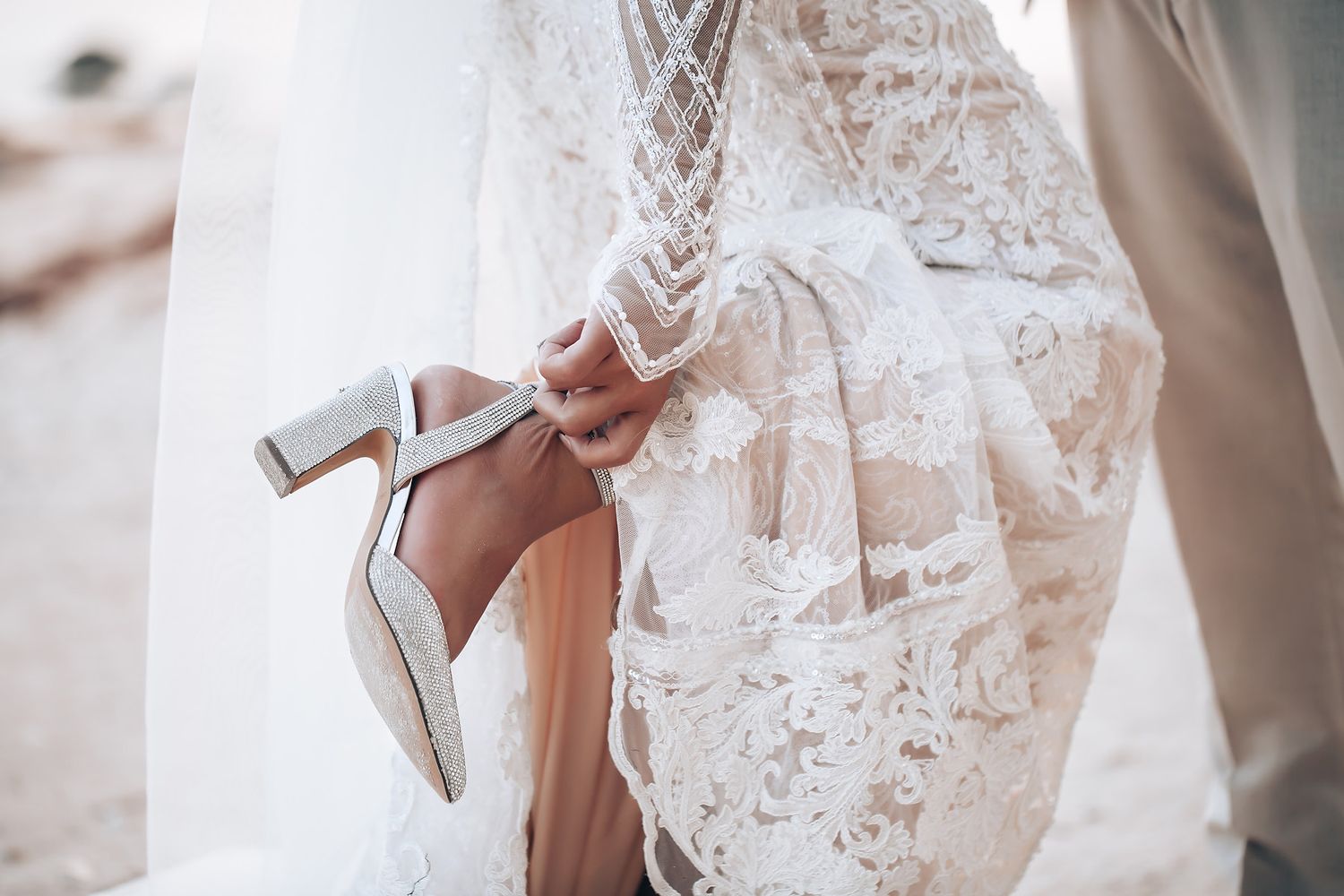 Bride in a delicate white lace wedding dress gets ready while holding elegant silver heels.