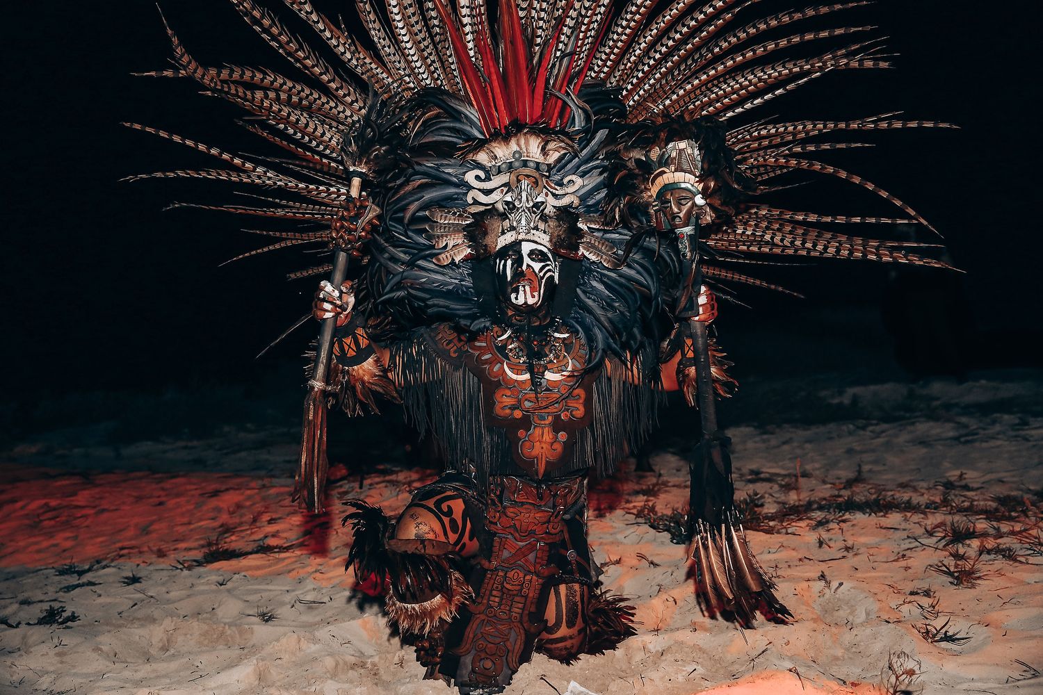 A dramatic figure in an Aztec ceremonial headdress and traditional costume poses against a dark background with red lighting effects.