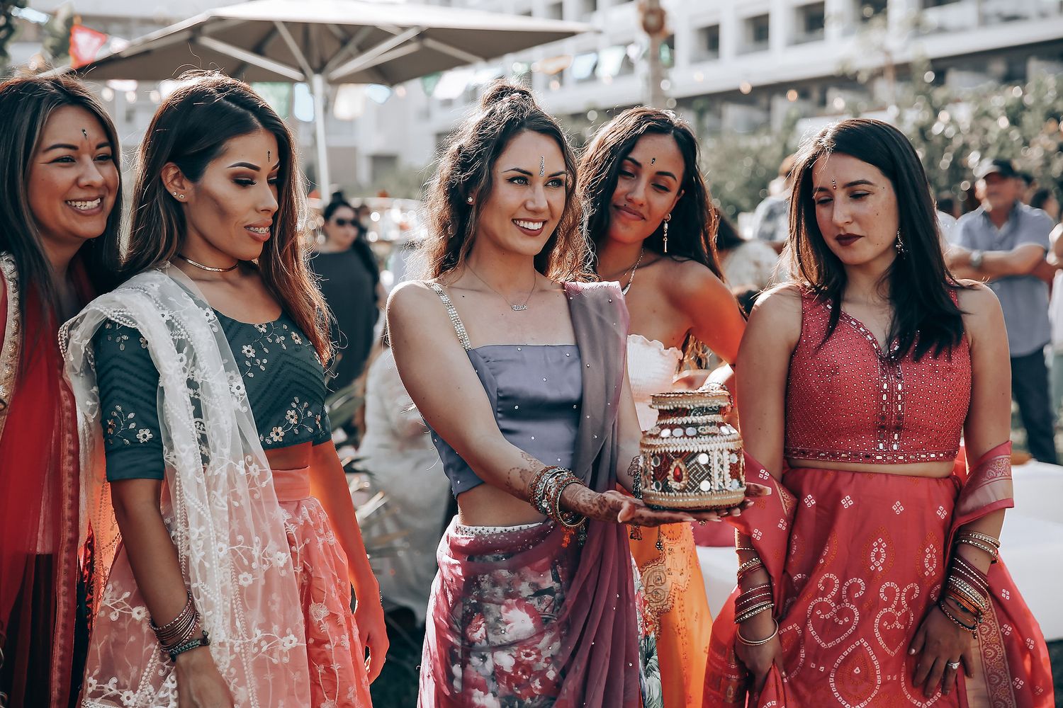 A group wearing traditional red and white Indian wedding attire poses together at an outdoor celebration.