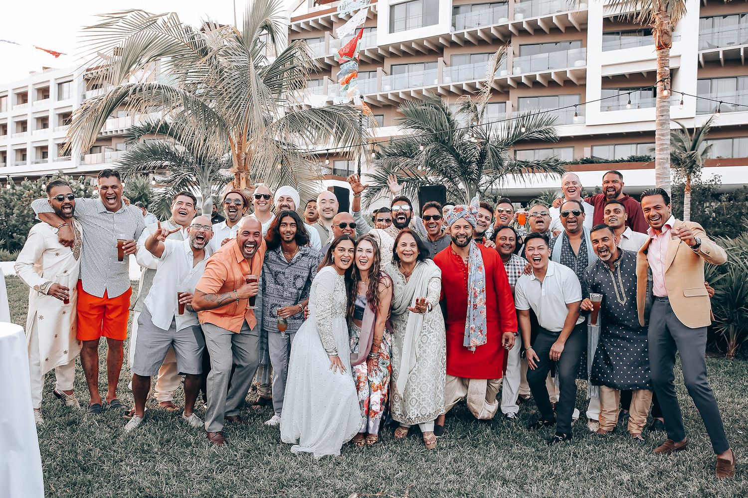 A large group of wedding guests pose together on a lawn in front of a resort hotel with palm trees.
