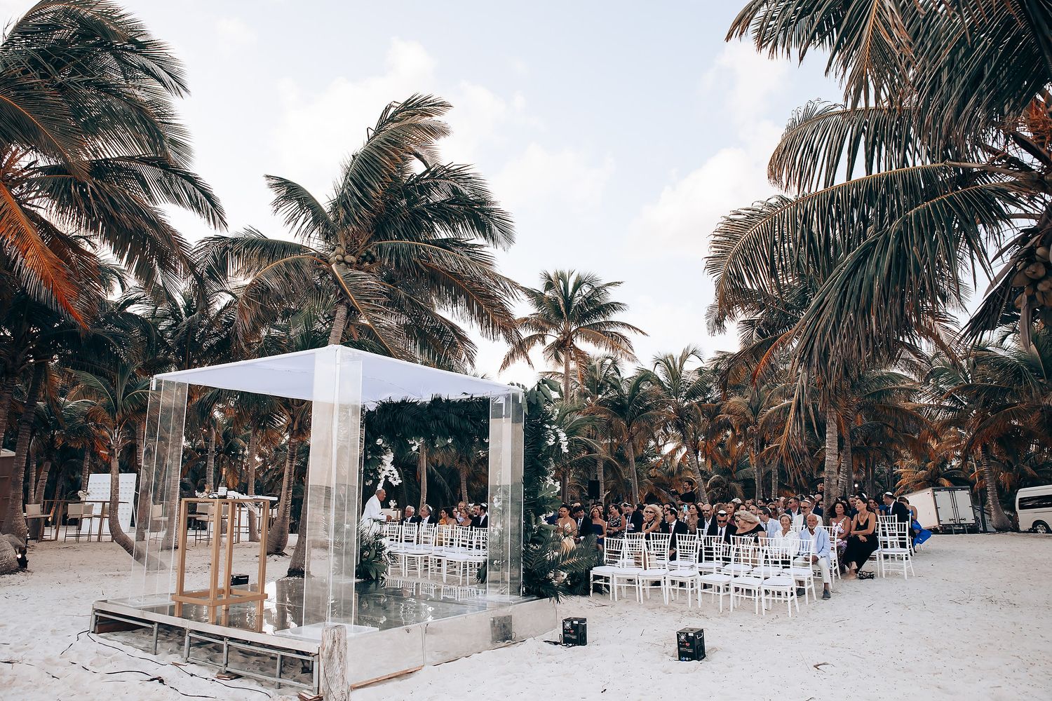 Panoramic view of a beach wedding setup with palm trees and white decor along the shoreline.