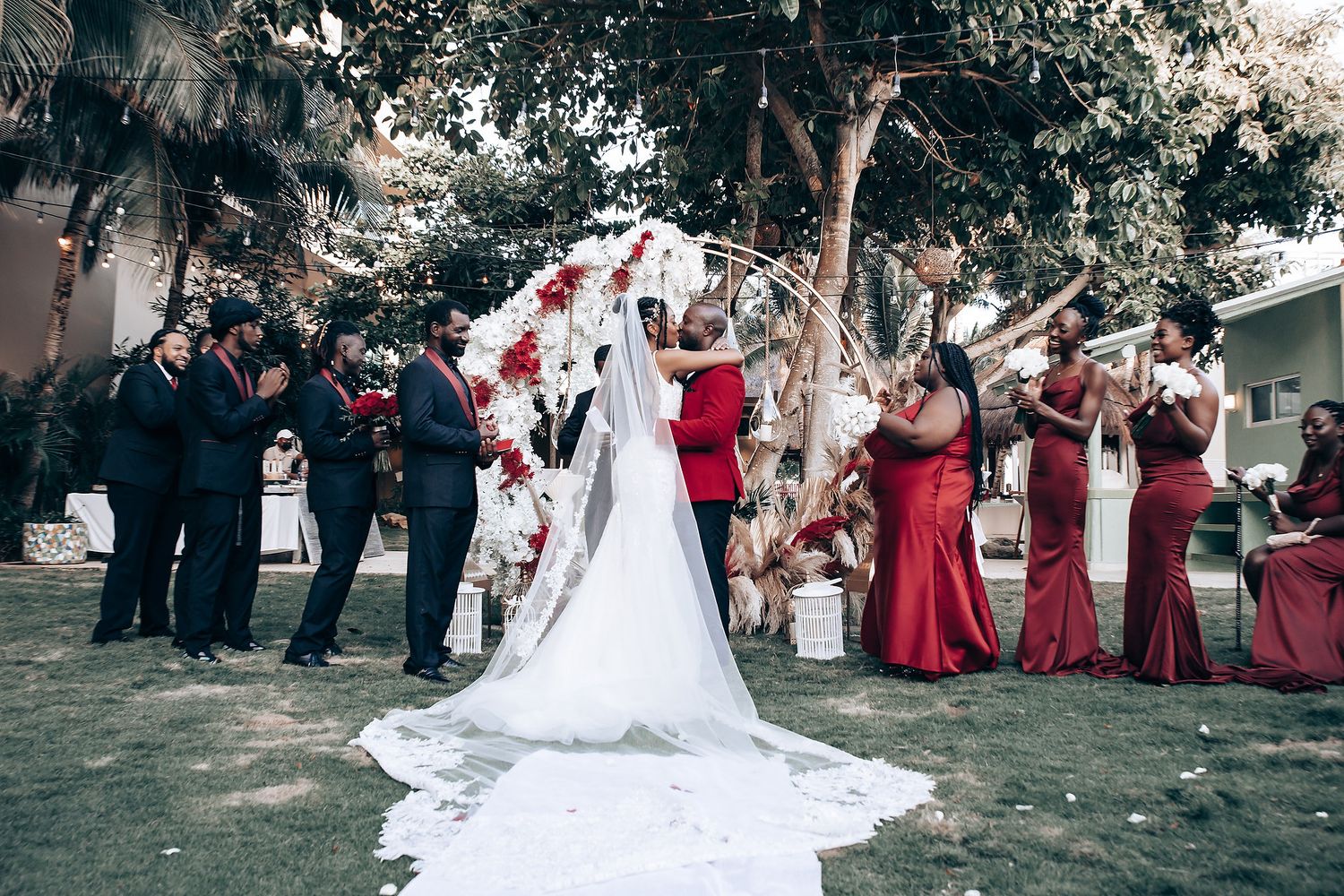 Wedding party in formal attire with bridesmaids in red dresses and groomsmen in black suits pose outdoors in garden setting.