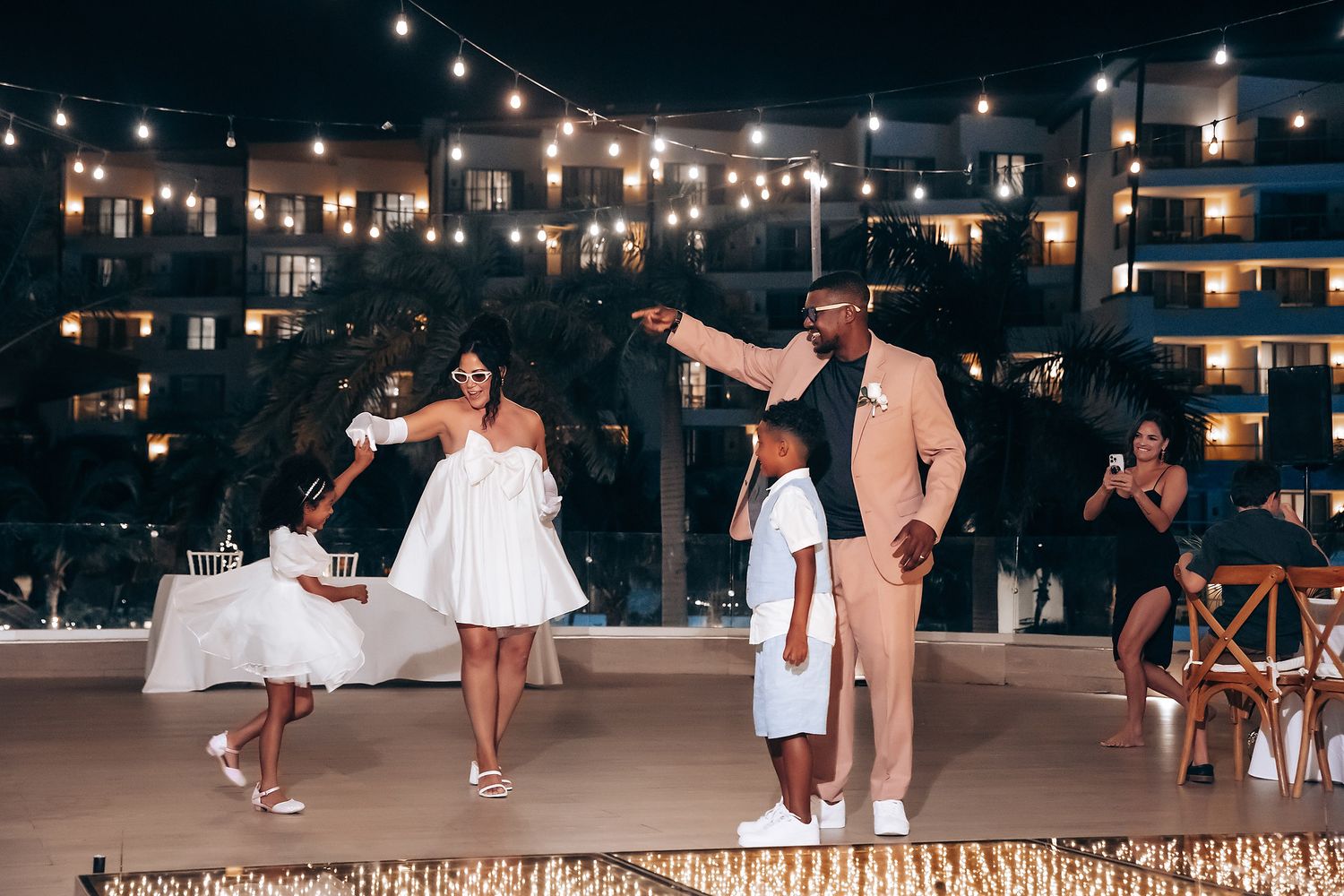Guests dance and celebrate on a beachfront patio under twinkling string lights at night.
