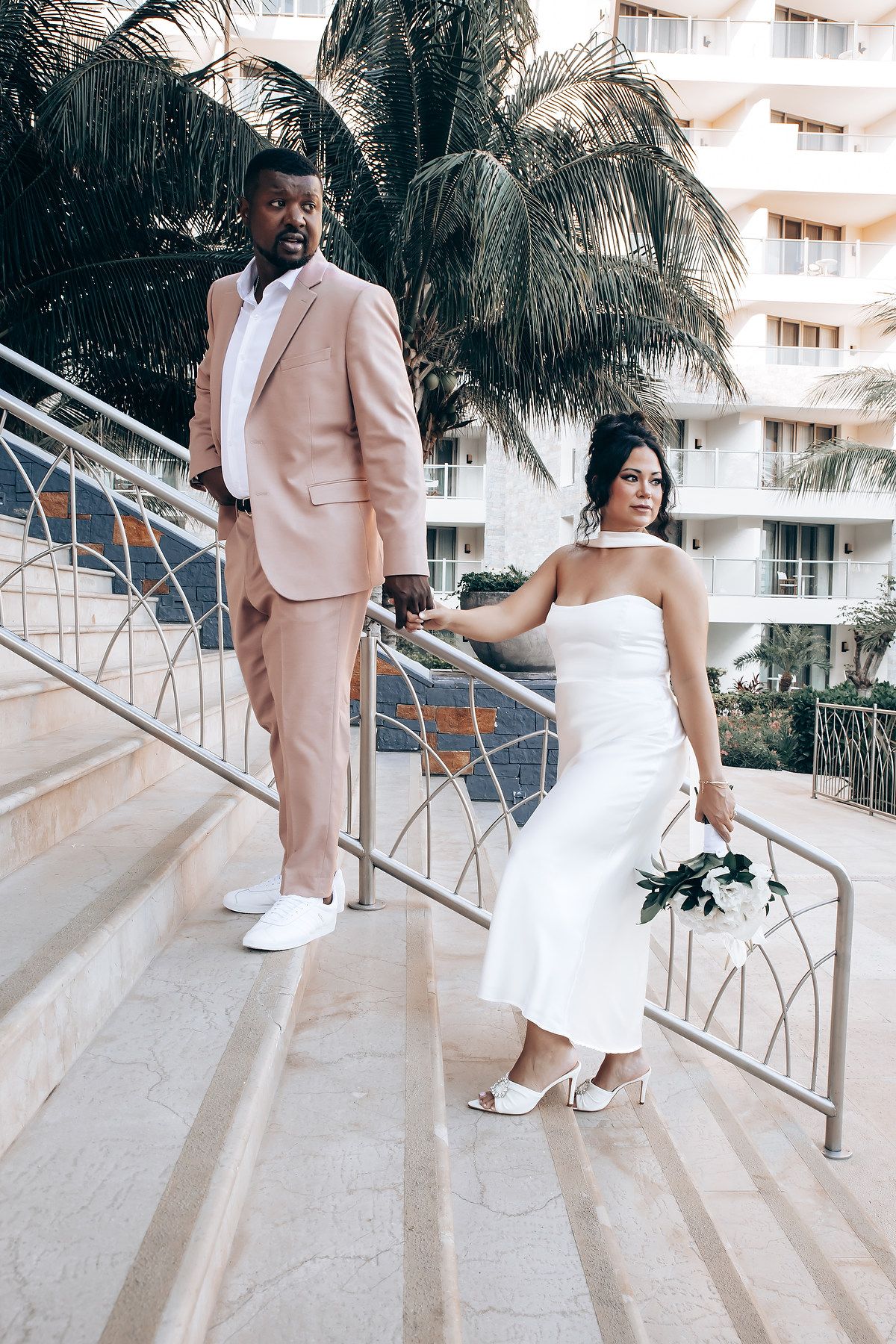 A couple poses on stairs with palm trees and a hotel in the background, wearing formal attire in pink and white.