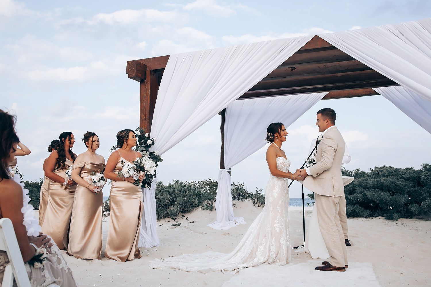 Wedding party poses on a sunny beach with flowing white curtains and beige attire creating a romantic atmosphere.
