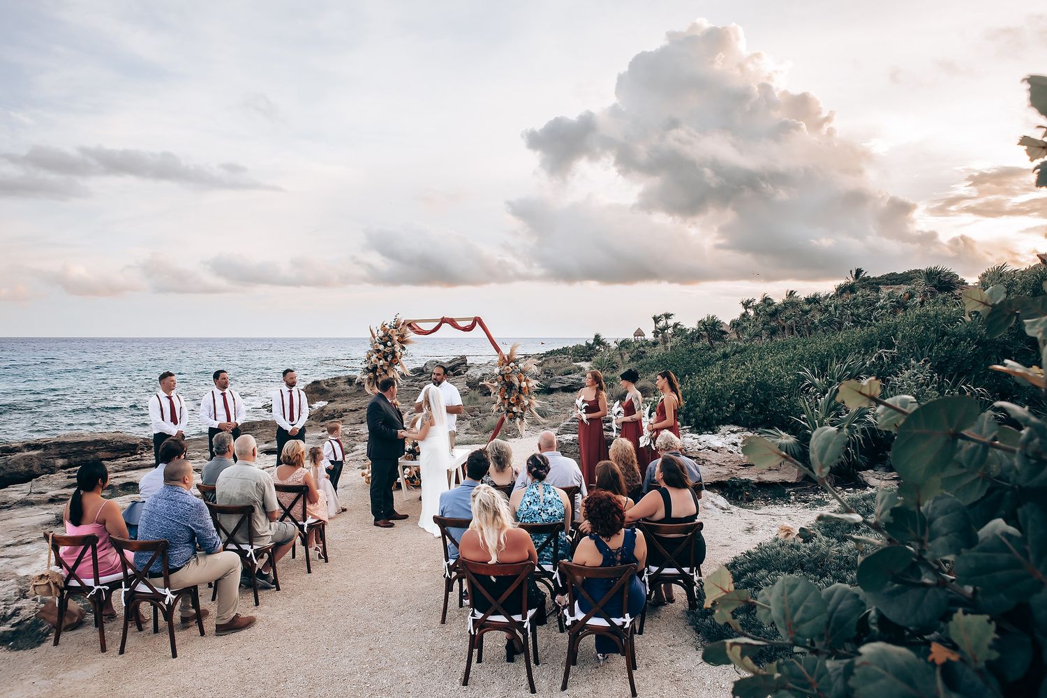 Guests gather for a sunset beach wedding ceremony with an arch decorated in tropical flowers on a rocky Hawaiian shore.