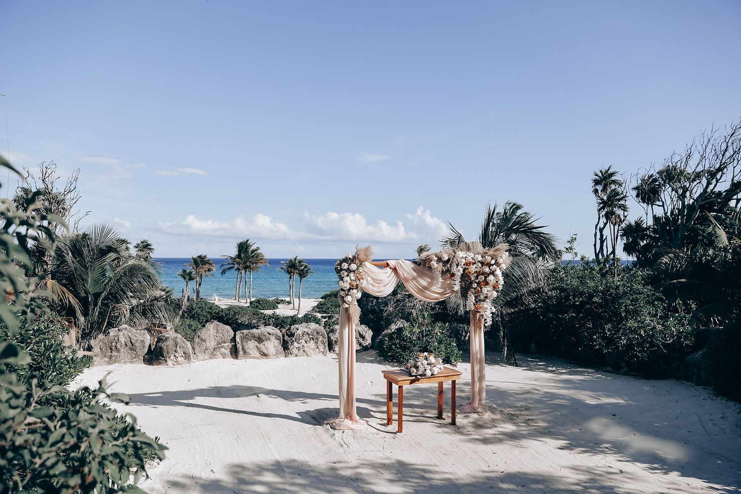 A wooden wedding arch with floral decorations overlooks a tropical beach with palm trees and turquoise ocean waters.