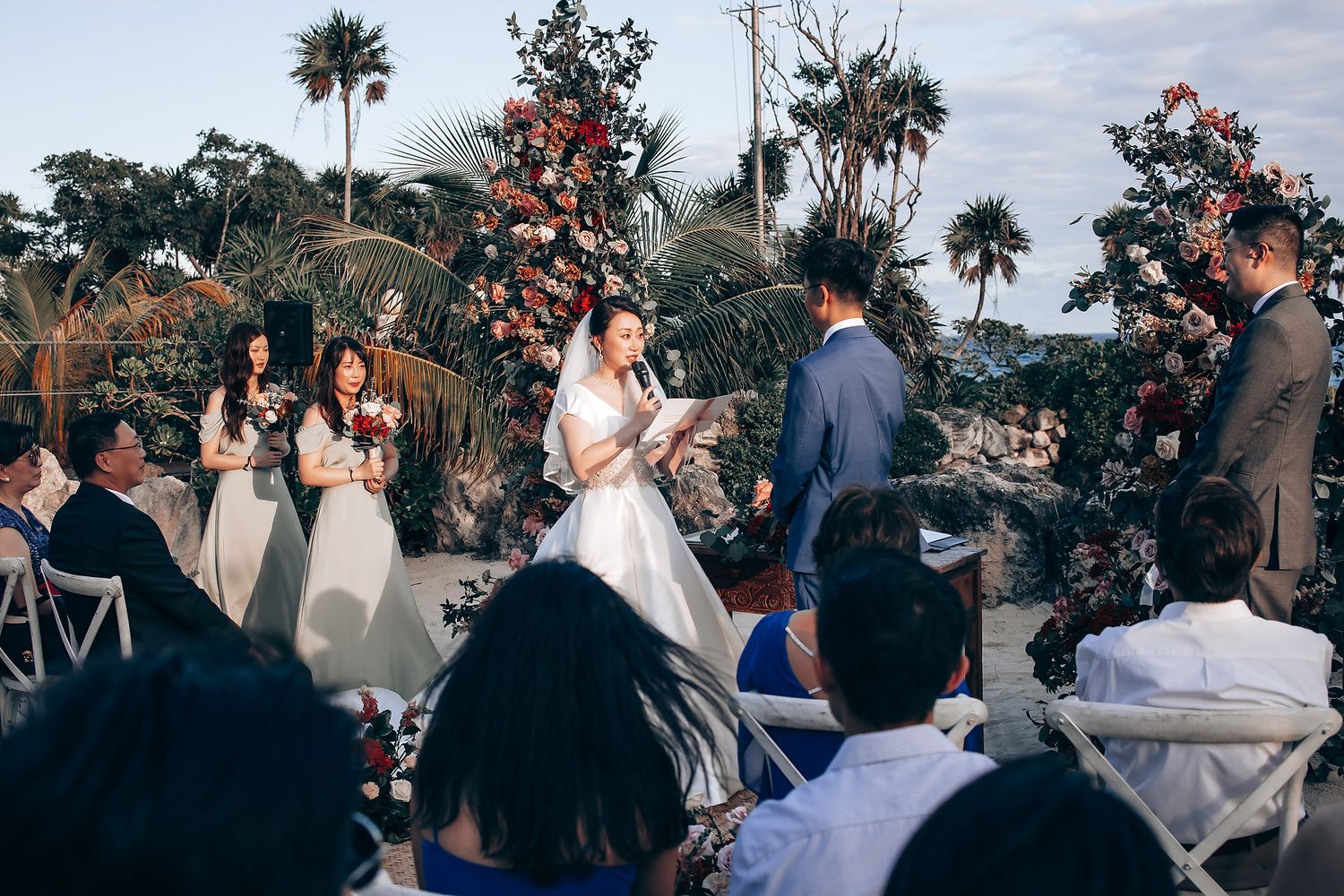 Wedding ceremony gathering with palm trees and decorative flowers in background.