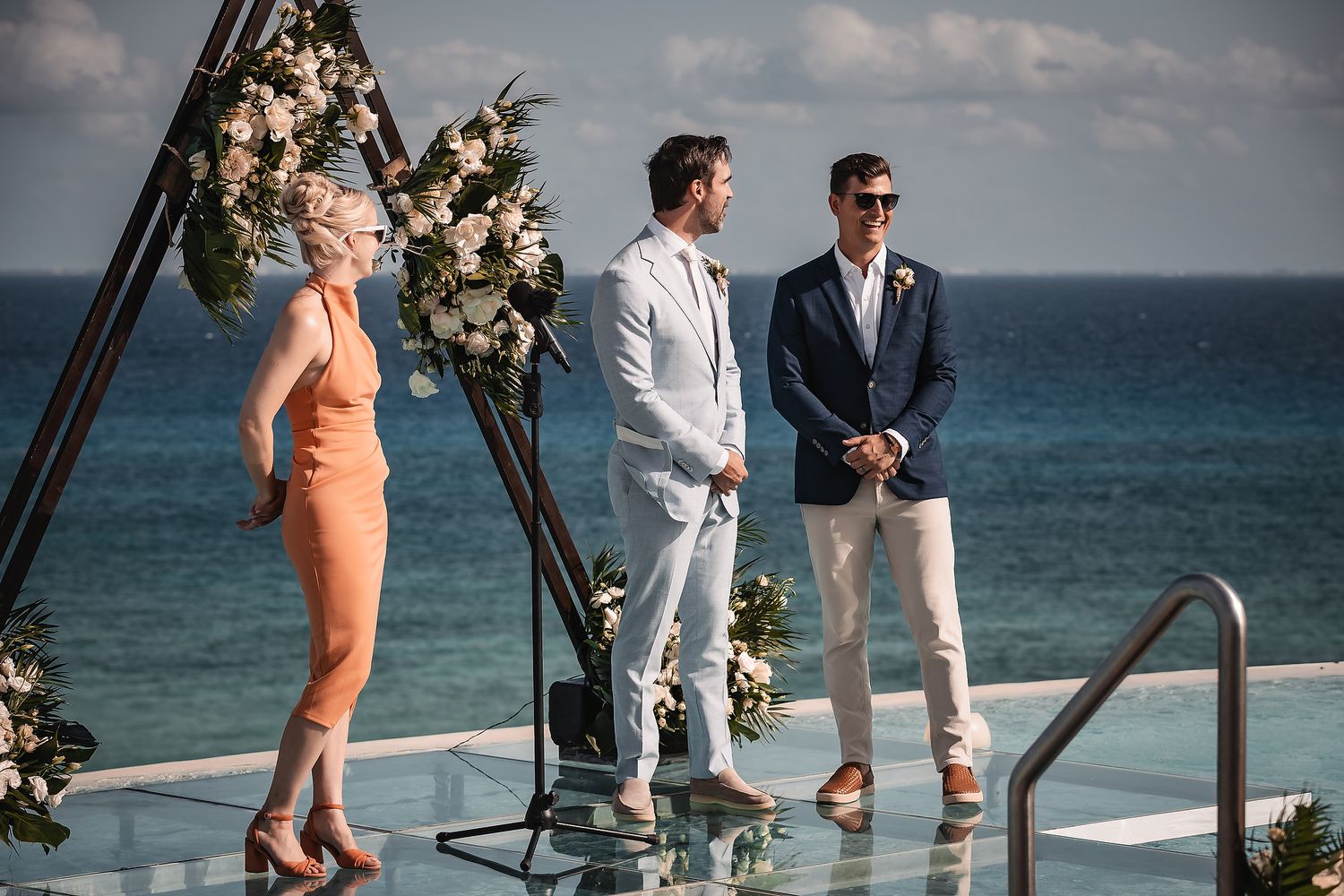 Wedding ceremony participants gather on an oceanfront deck under blue skies.