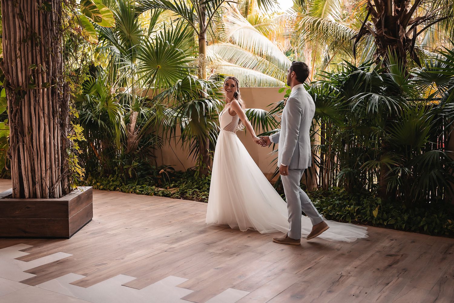 A couple walks together in a tropical garden setting with palm trees and natural light.