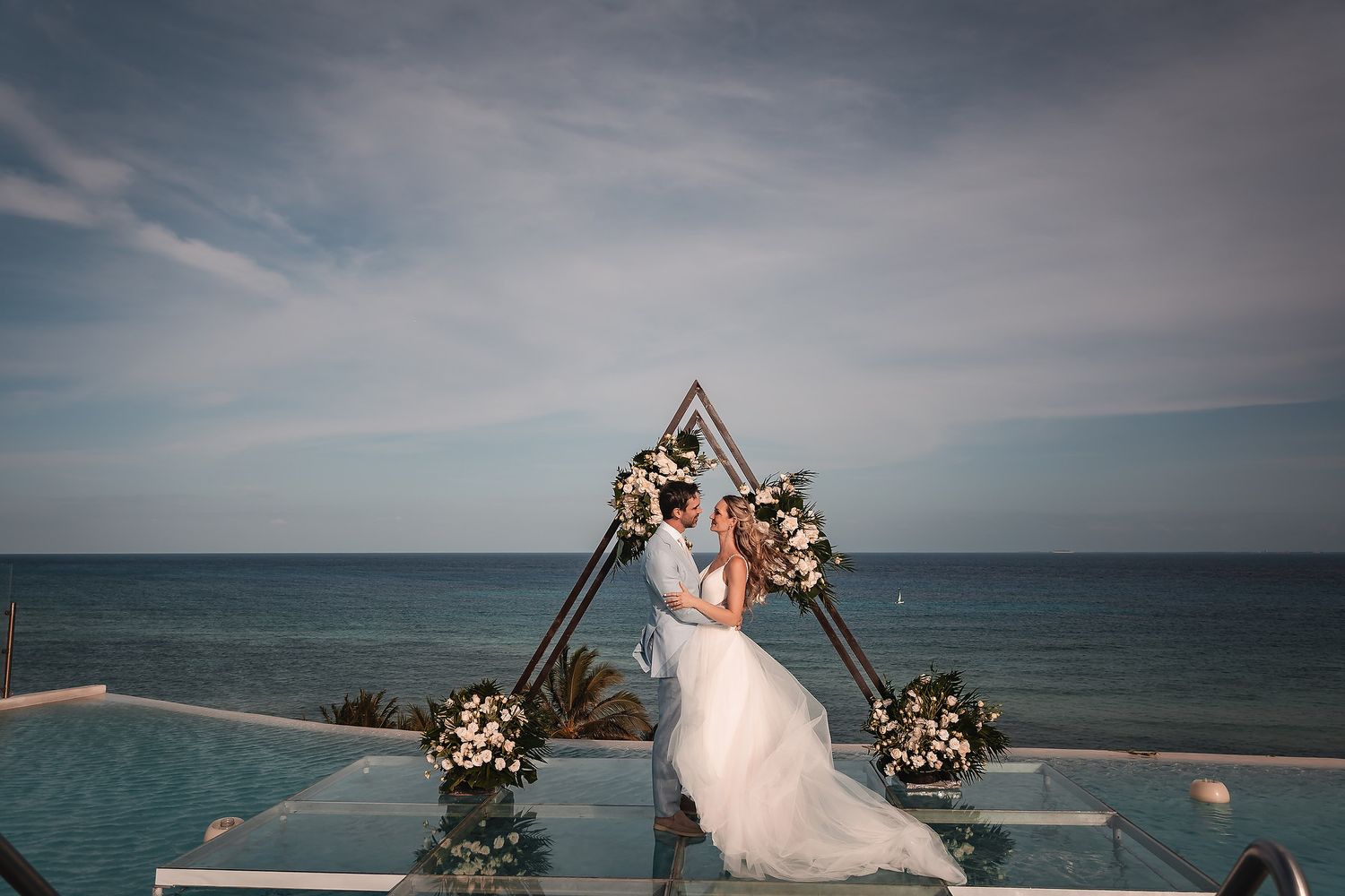 Scenic beach wedding setup with geometric arch decorated in blush roses overlooking the ocean.