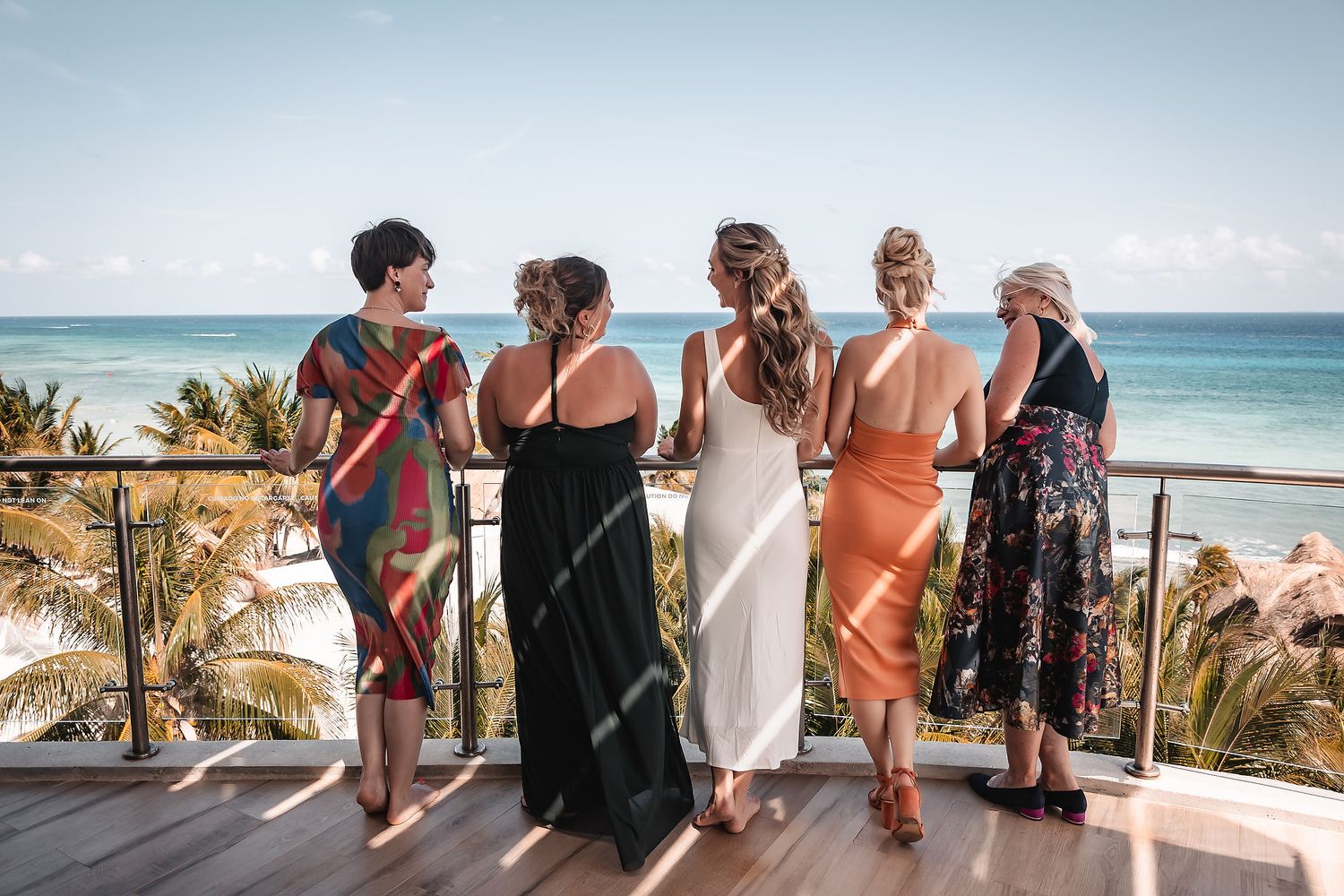 Wedding guests gather on a sunny beachfront balcony with palm trees and ocean views.