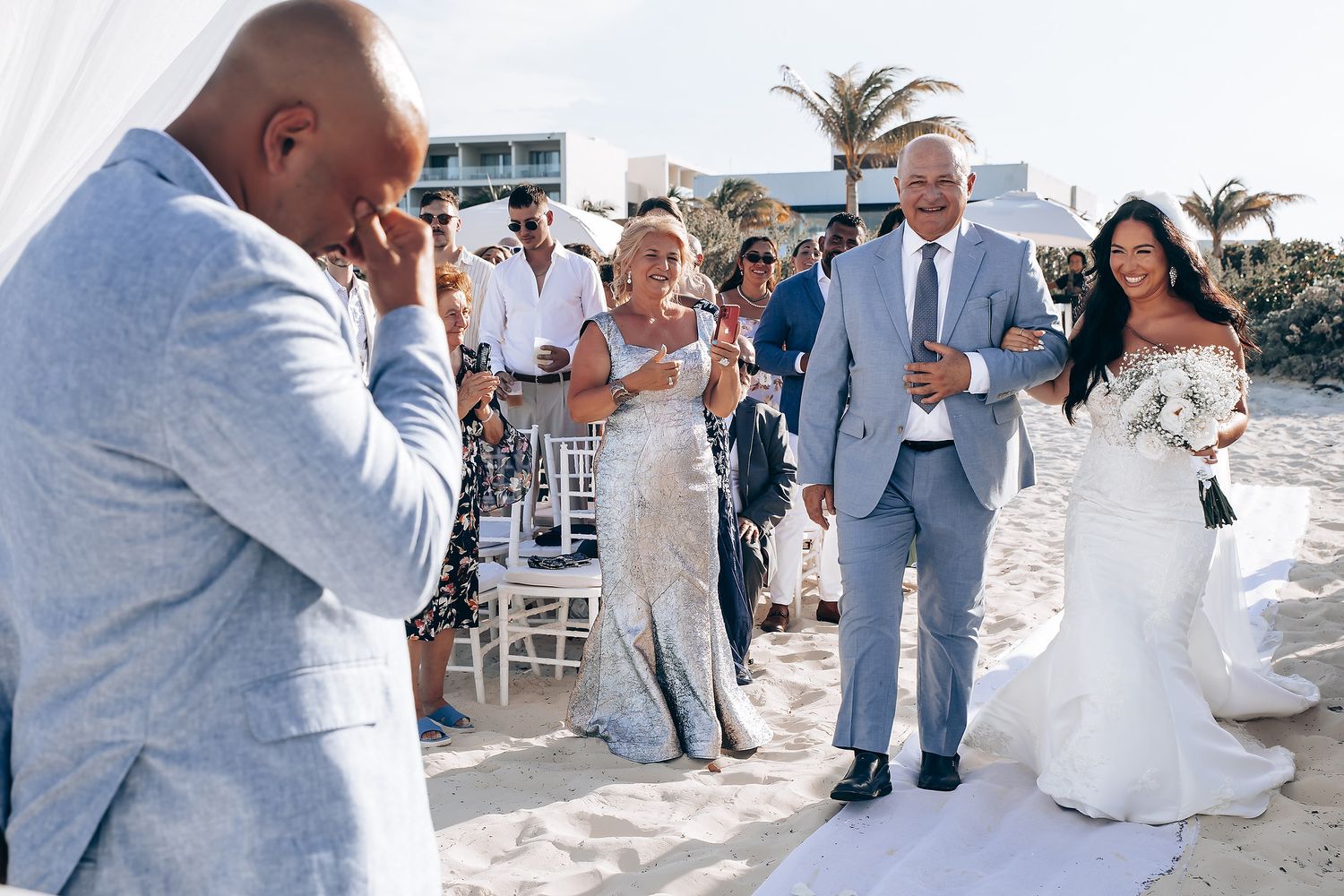 Wedding celebration on a beach with guests in light blue and white attire dancing near palm trees.