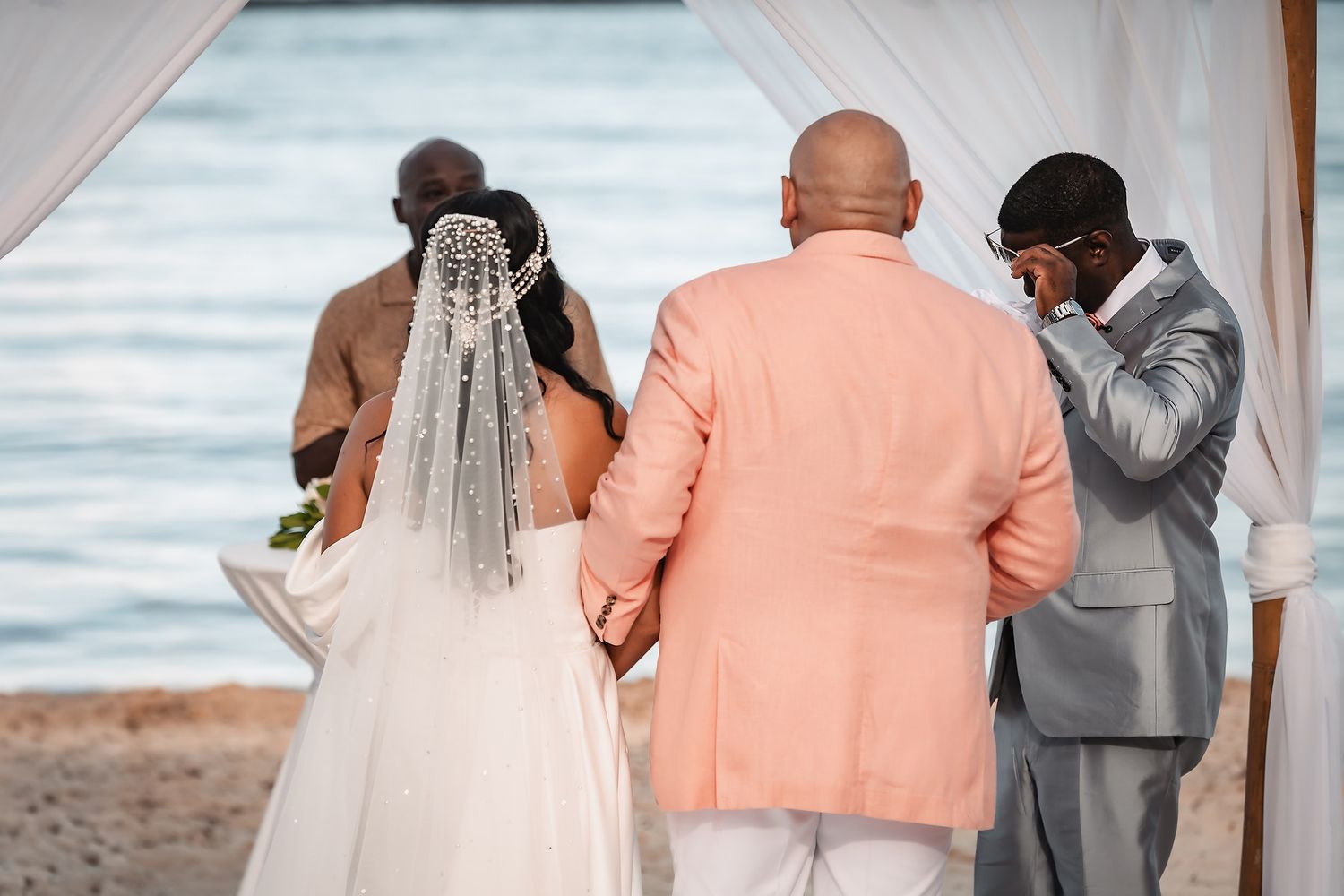 Wedding ceremony on a beach with attendees in formal attire against an ocean backdrop.