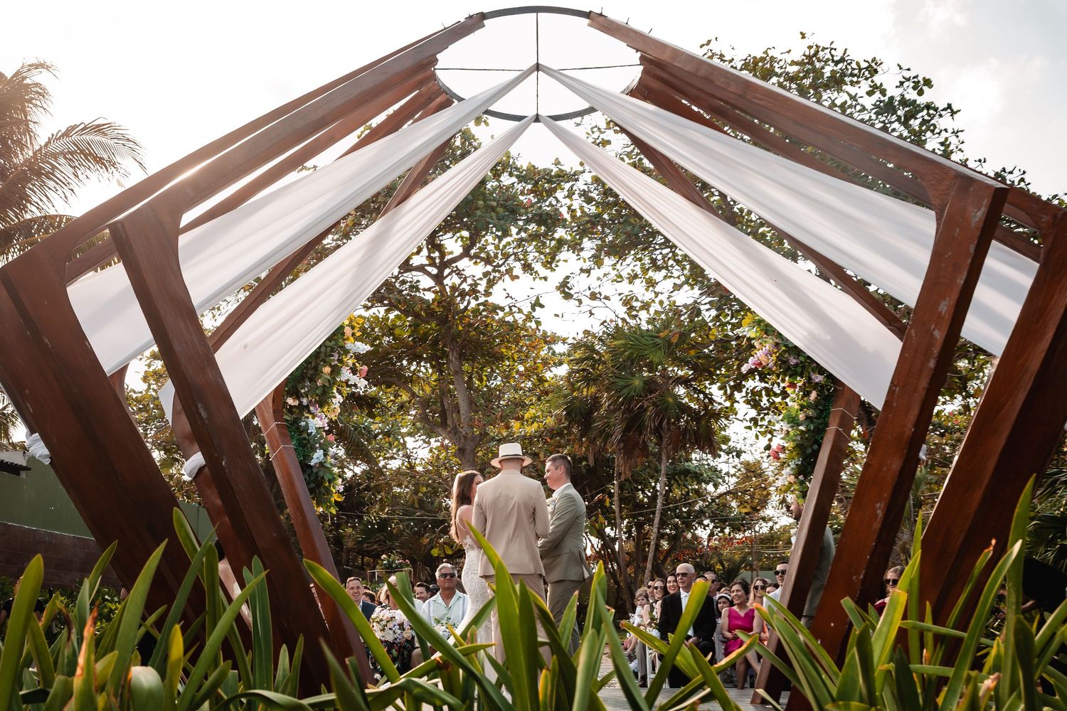 Panoramic view of wooden wedding arch with white draping and floral decorations against tropical backdrop.