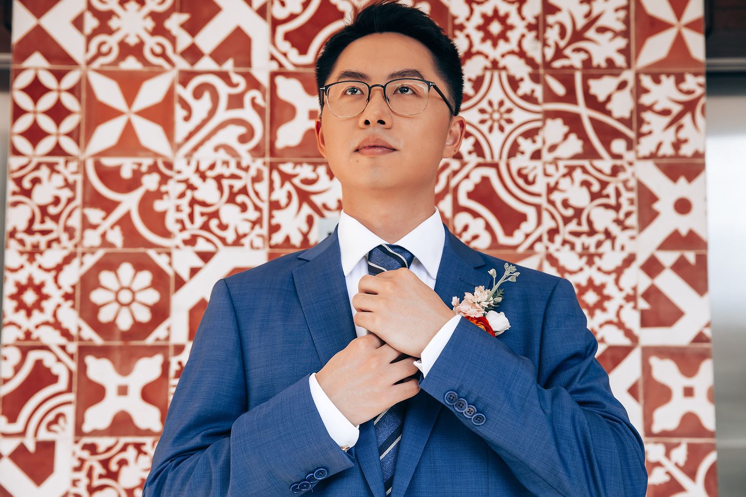 A groom in a navy blue suit and glasses adjusts his tie against a decorative red and white patterned backdrop.