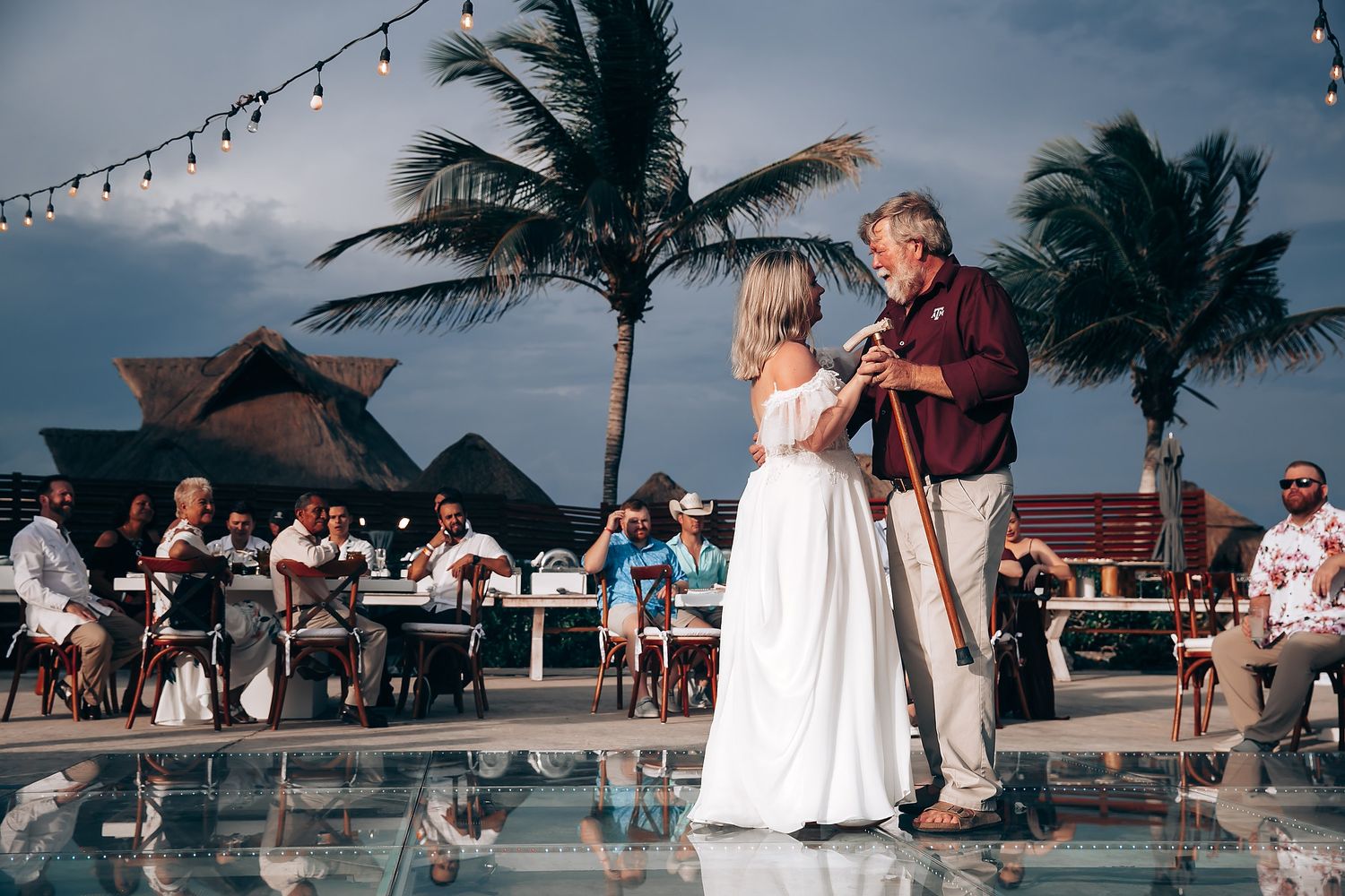 Dancing scene at outdoor wedding reception with string lights and palm trees at dusk.