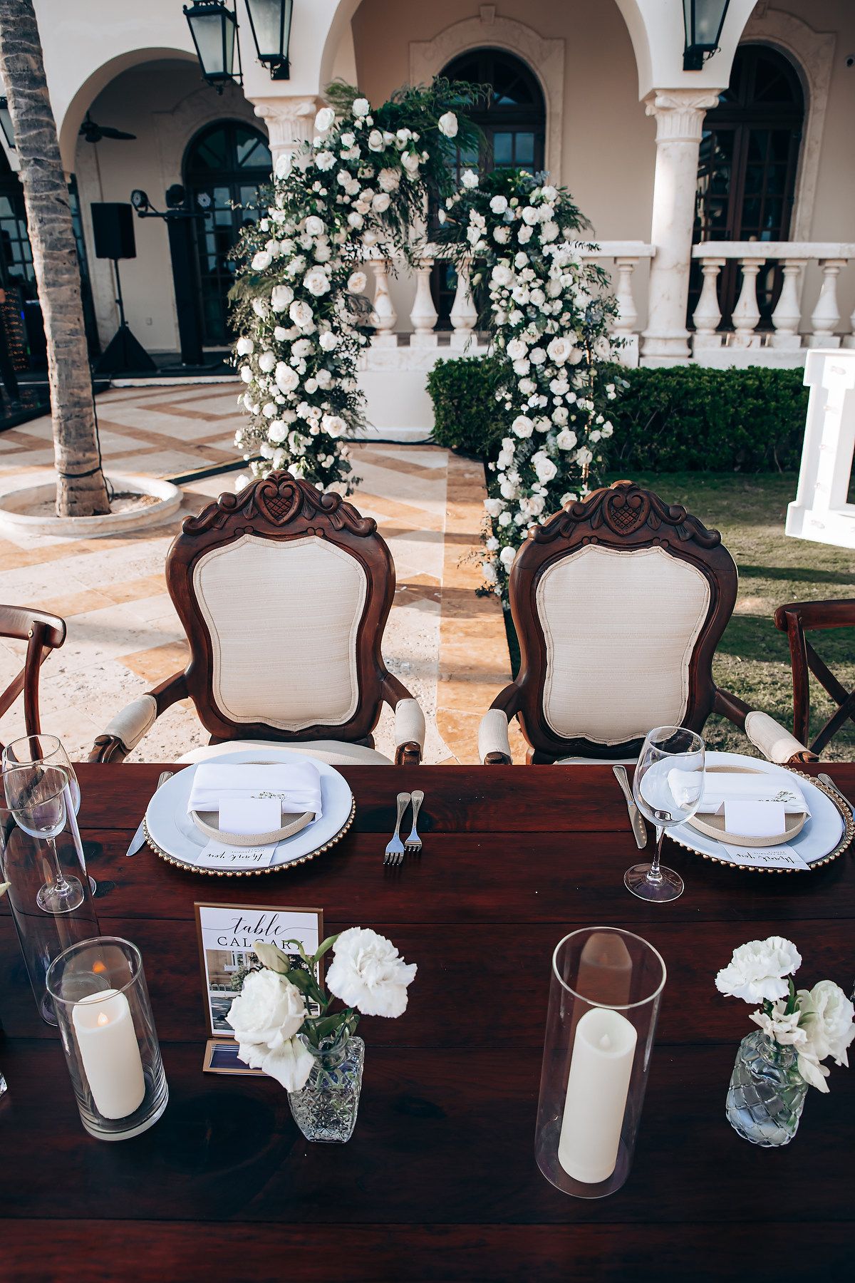 Elegant wedding reception table setup with ornate white chairs, place settings, and floral arrangements in an outdoor venue.