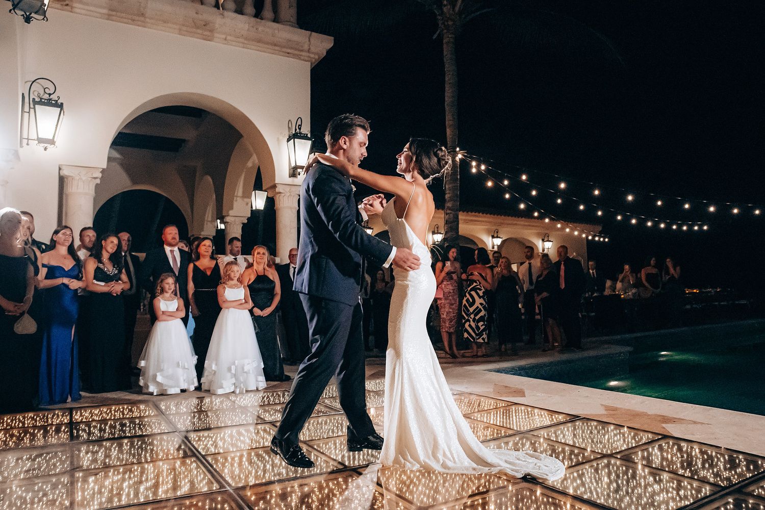 Wedding couple and guests dancing together on an illuminated outdoor patio with string lights overhead.