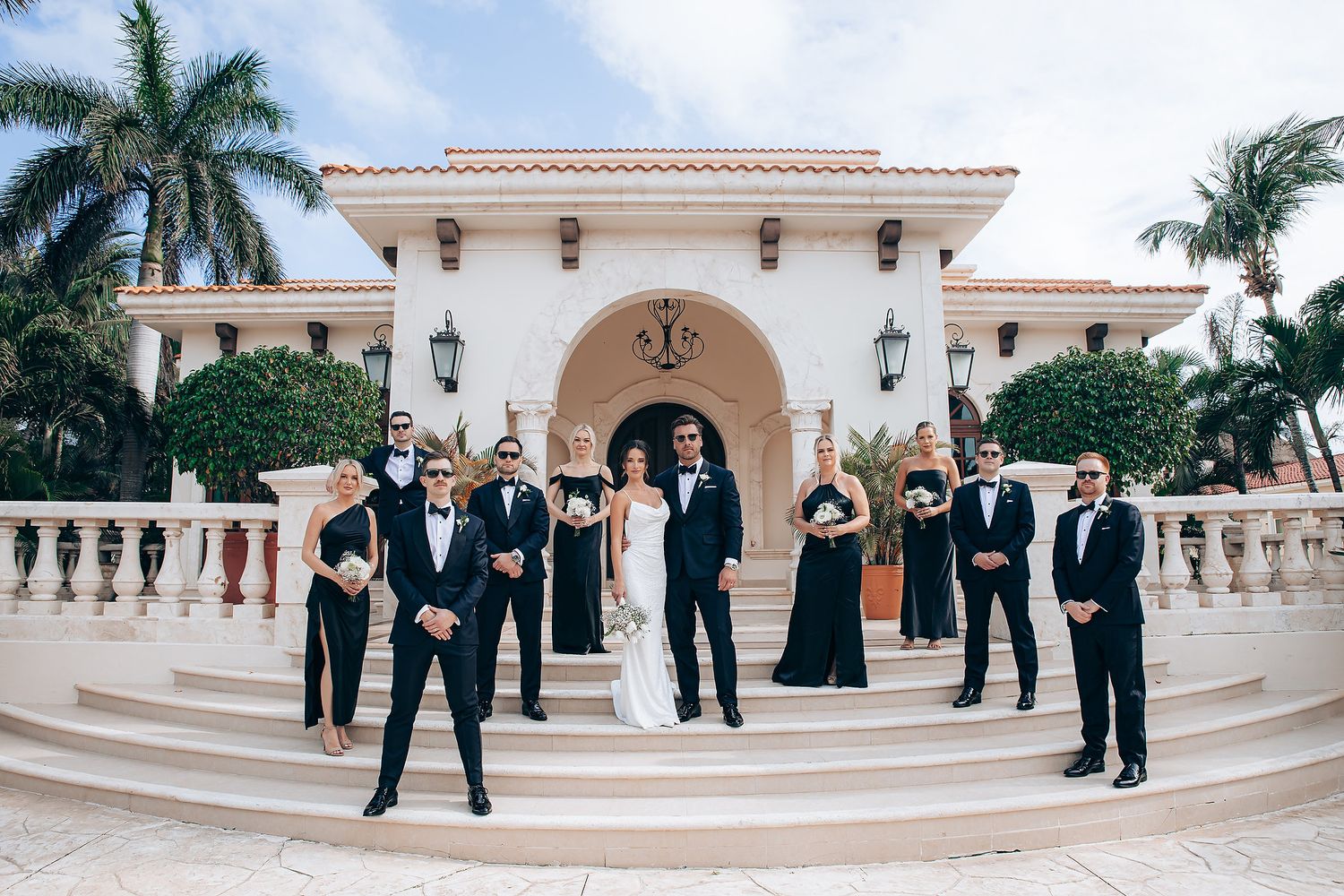 Wedding party members in formal attire pose on an outdoor terrace with palm trees and Mediterranean architecture.