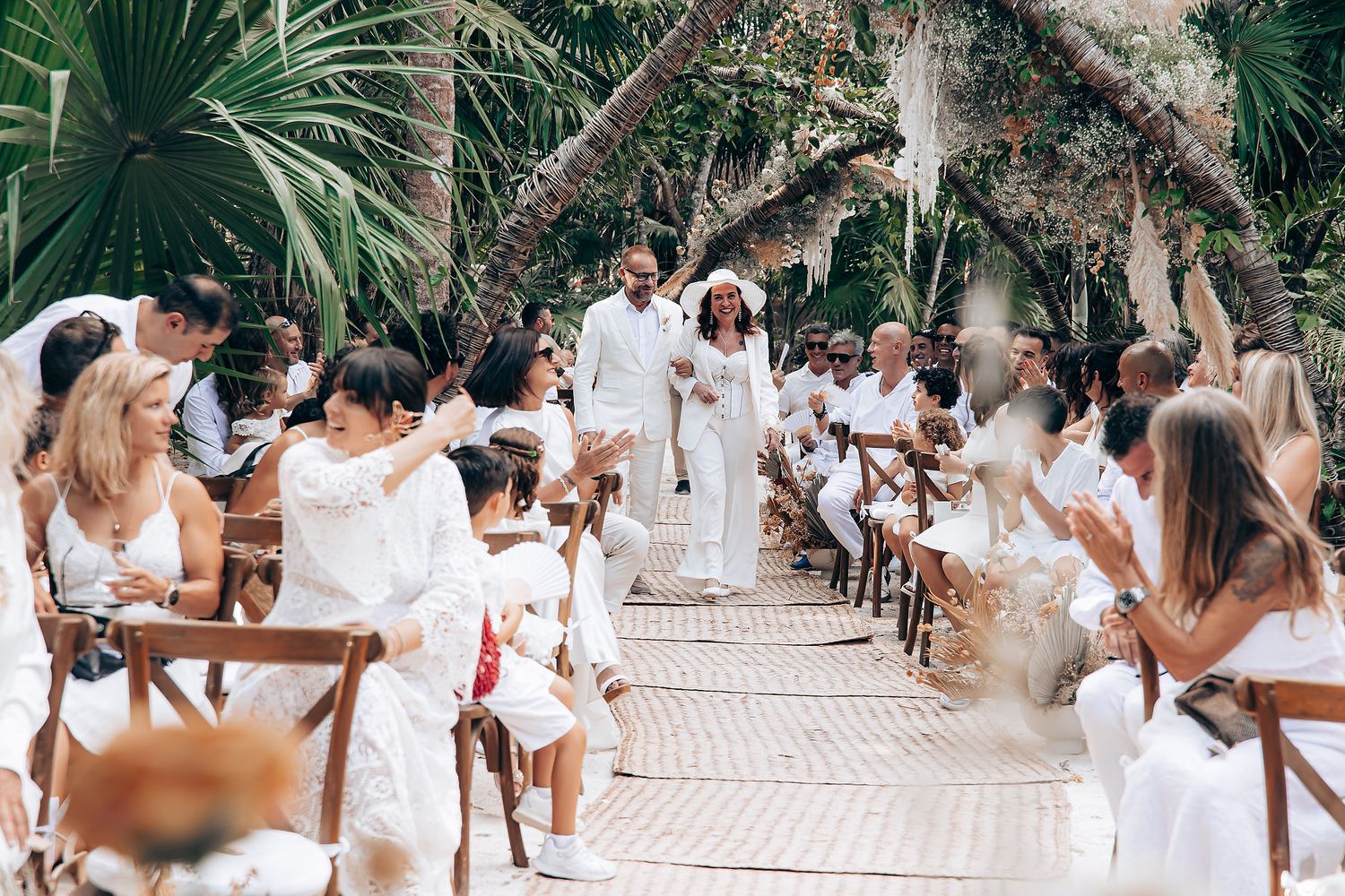 Wedding guests in white attire gather at long outdoor tables under tropical palm trees for a reception celebration.
