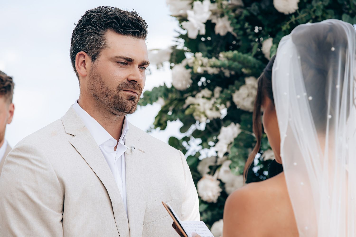 A couple shares a tender moment during their outdoor wedding ceremony surrounded by white floral decorations.