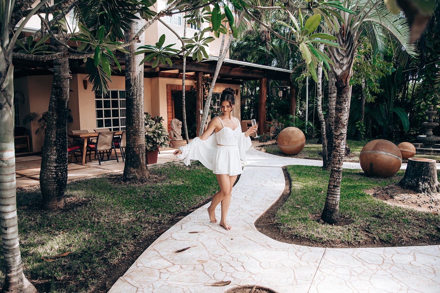 A person walks along a winding concrete path surrounded by palm trees and tropical landscaping in a resort setting.