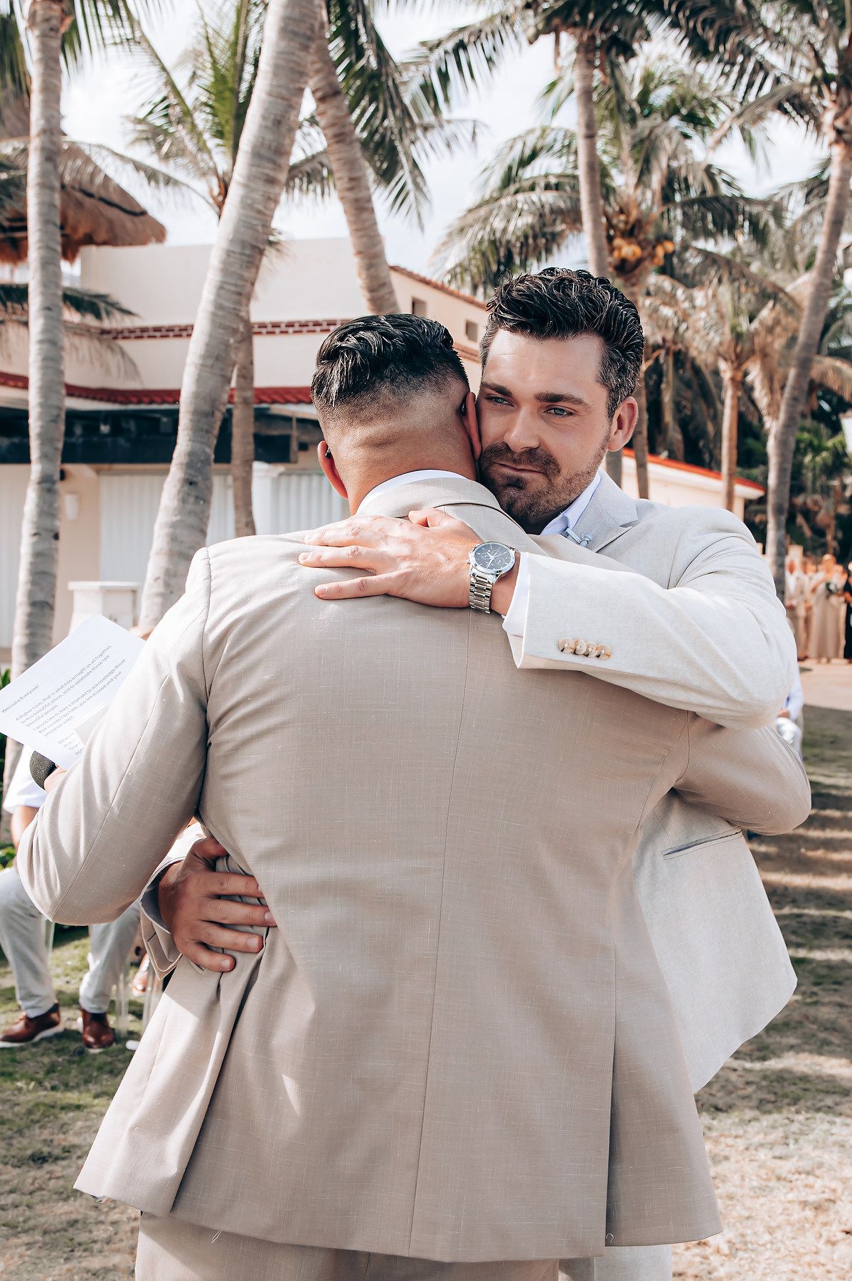 Groomsmen in beige suits share emotional embraces under palm trees at a tropical wedding venue.