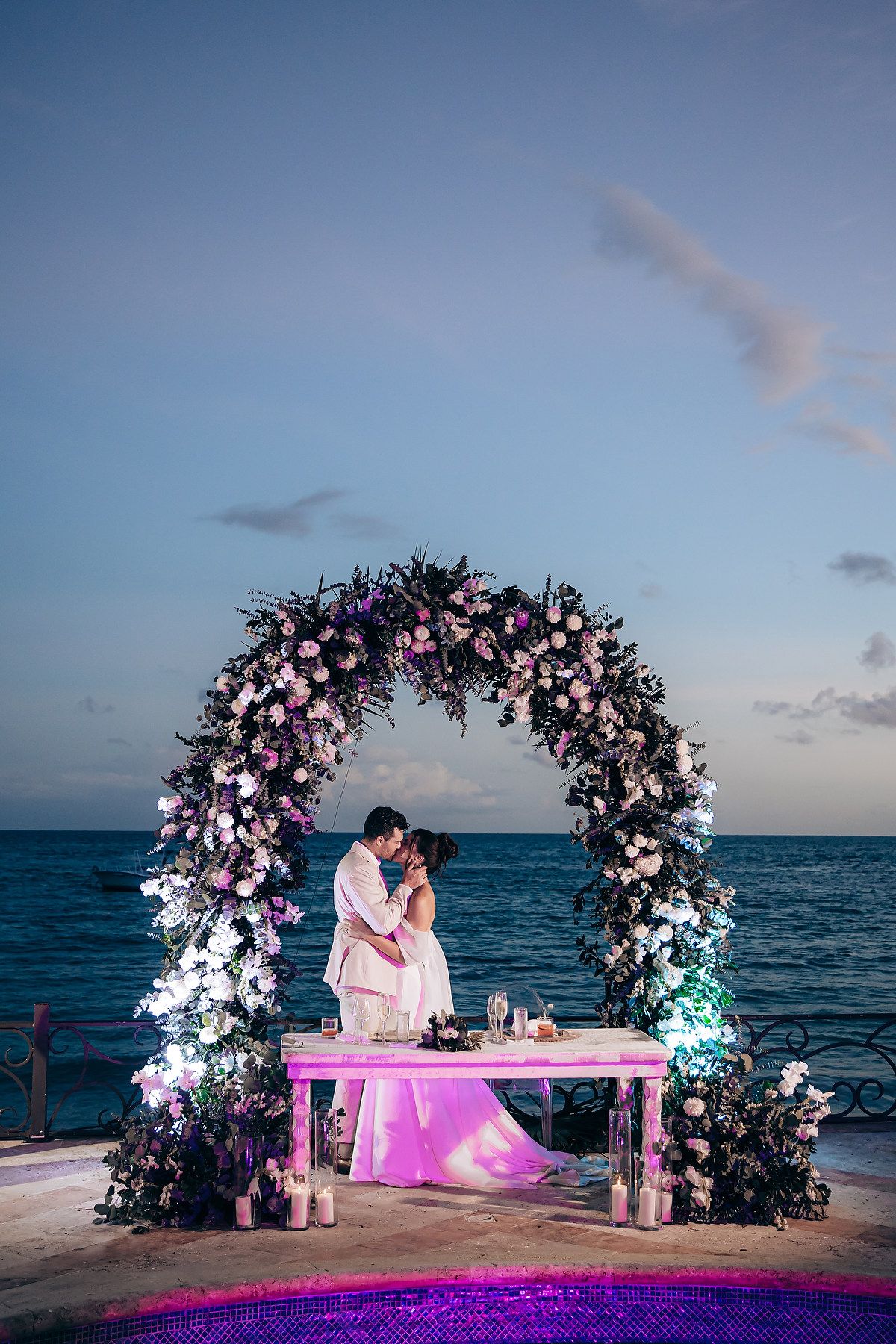Beachside wedding ceremony setup with illuminated floral arches and pink lighting against an ocean backdrop at dusk.