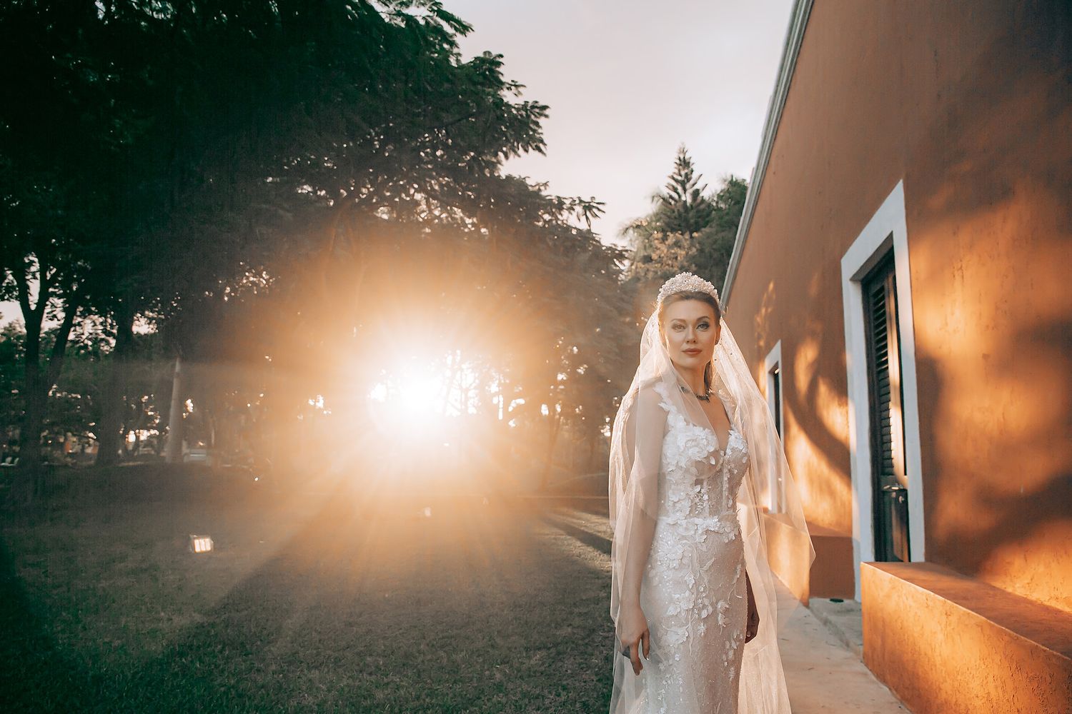 A person in a white lace wedding dress stands against a sunlit orange wall during golden hour.