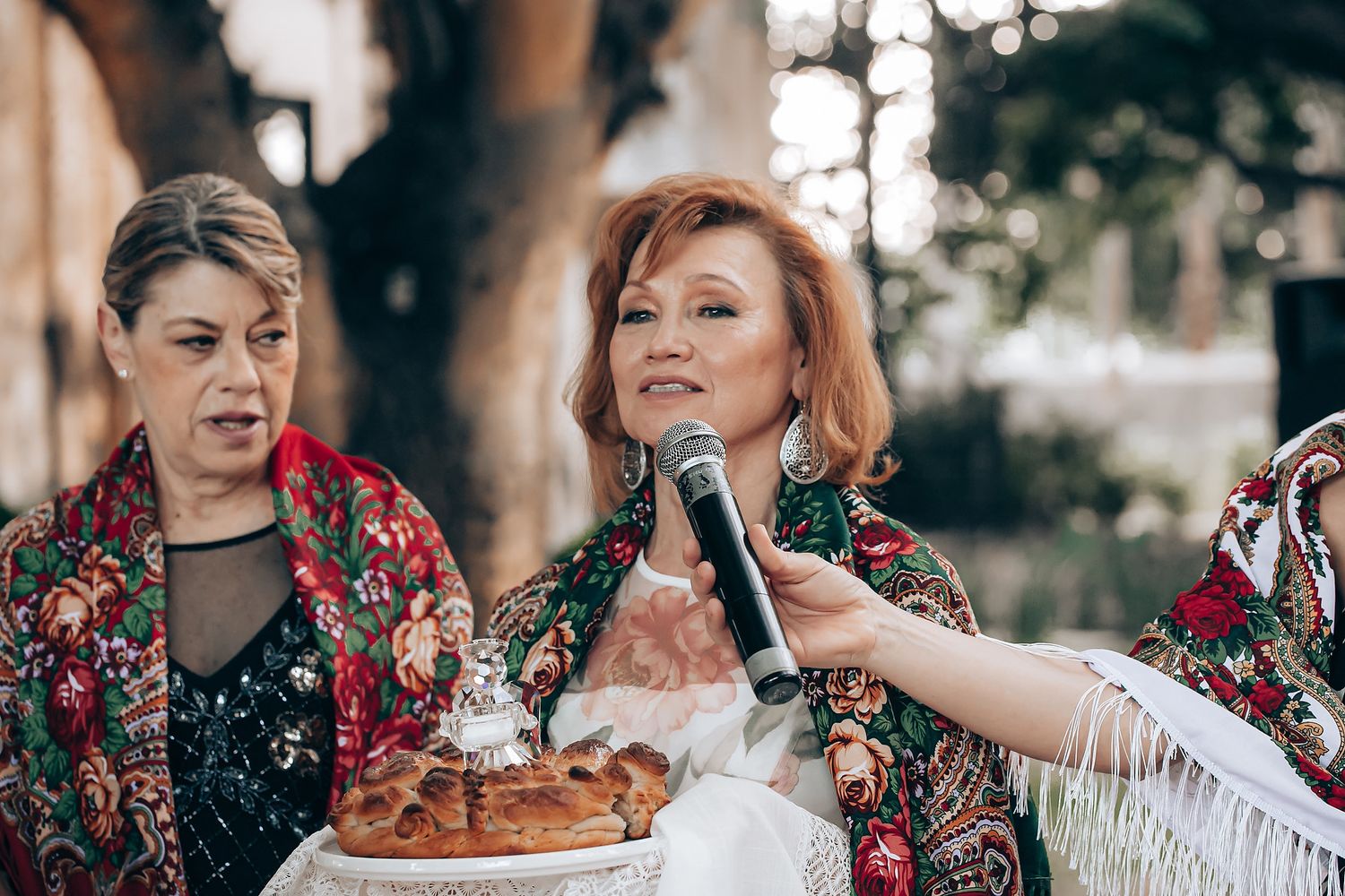Two people in floral dresses share a festive moment with cake and drinks at an outdoor celebration.