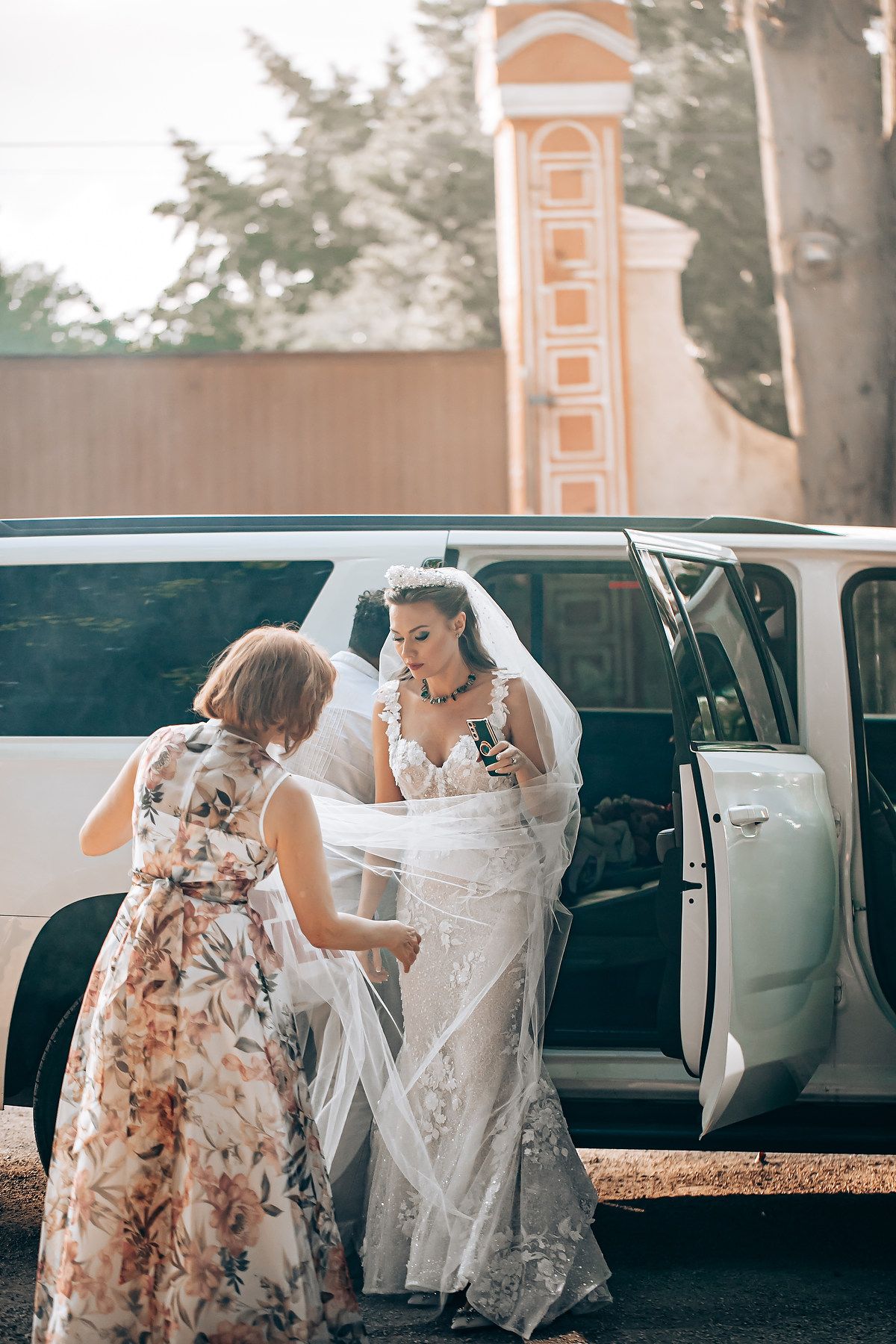 A bridesmaid in a floral dress helps the bride exit a black car before the wedding ceremony.