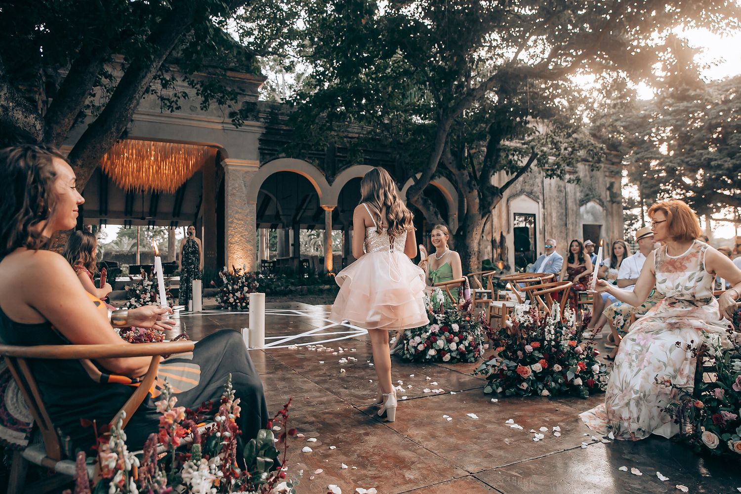 A romantic outdoor wedding ceremony with guests seated in white chairs under trees at sunset.