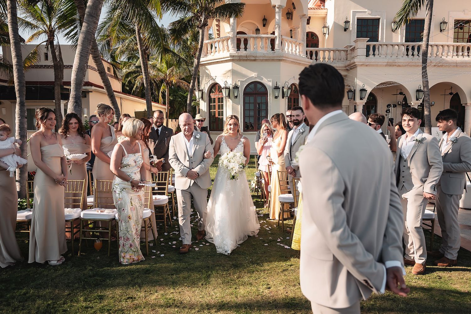 Wedding guests dressed in white gather at an elegant outdoor venue with palm trees and Spanish architecture.