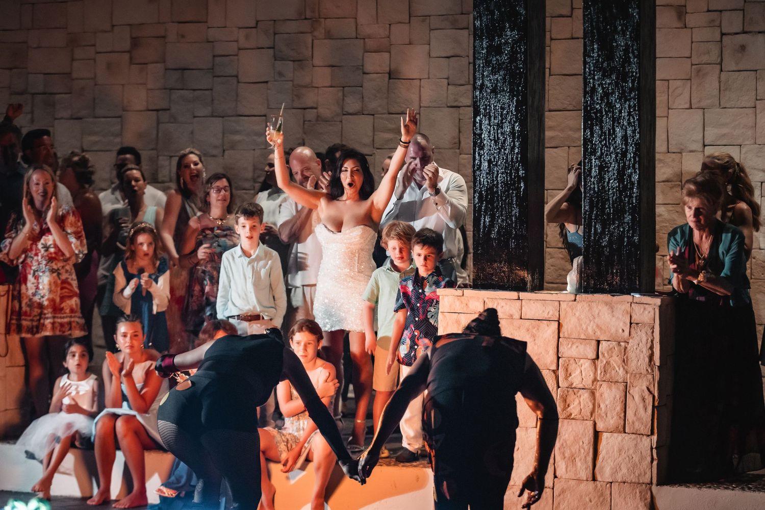 Crowd watching a lively performance in front of stone wall with dramatic lighting.