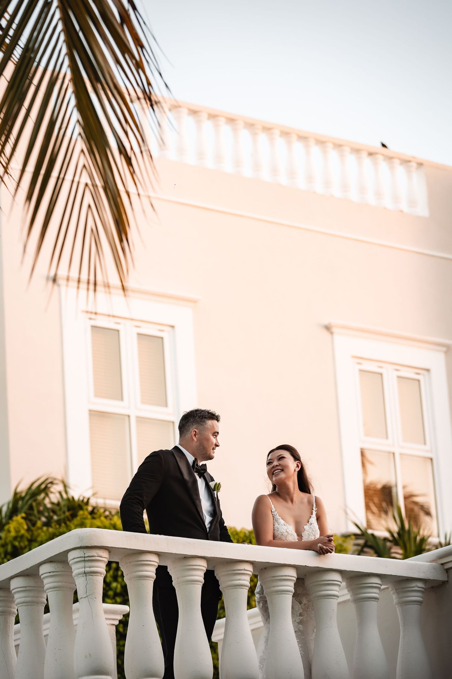 Newlyweds share a romantic moment on a white balcony with palm trees in the background.