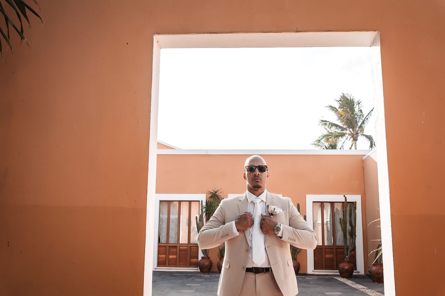 A person in a light colored suit adjusts their tie against a coral-colored wall with palm trees.