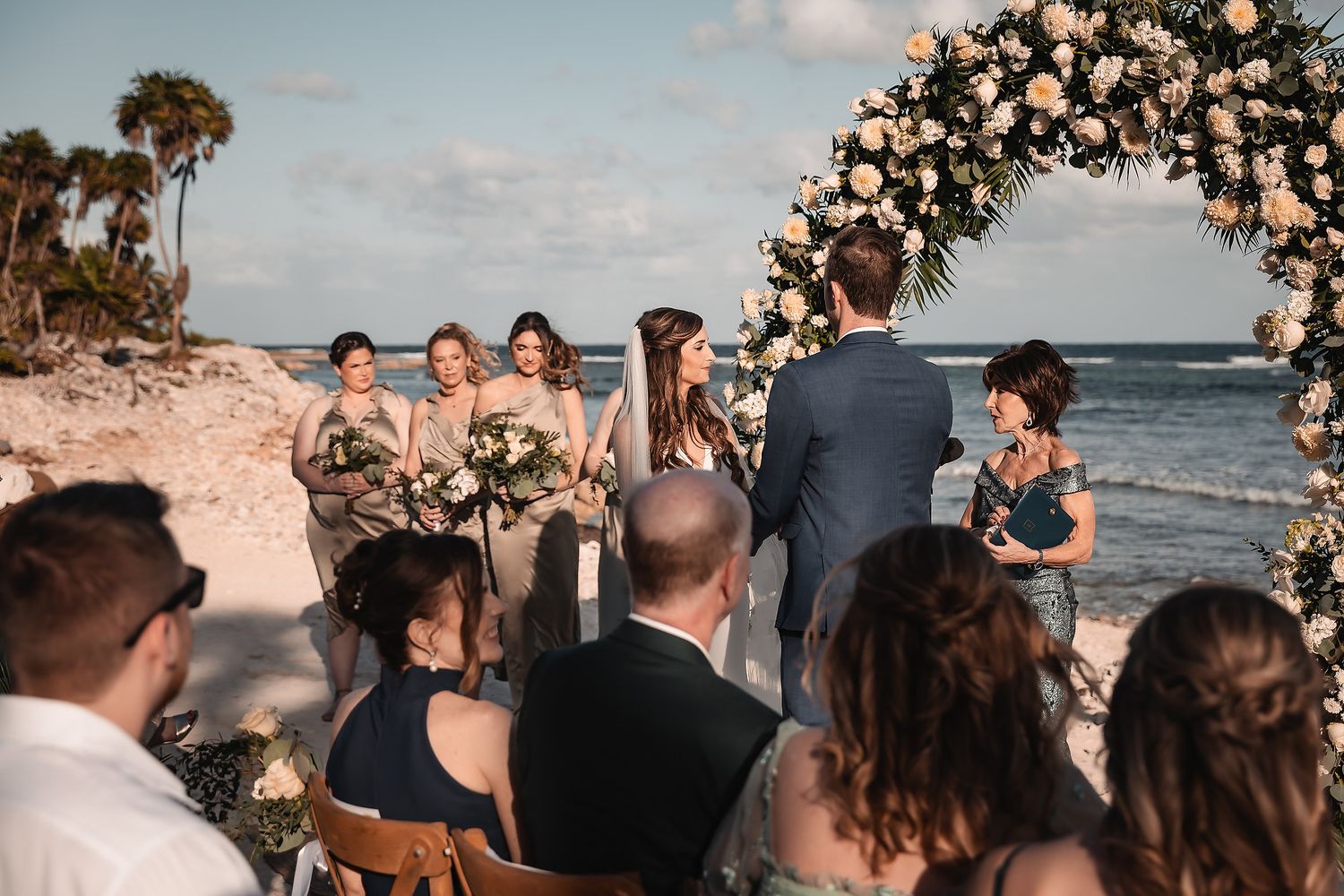 Intimate beach wedding ceremony with floral arch and guests gathered by the shoreline.